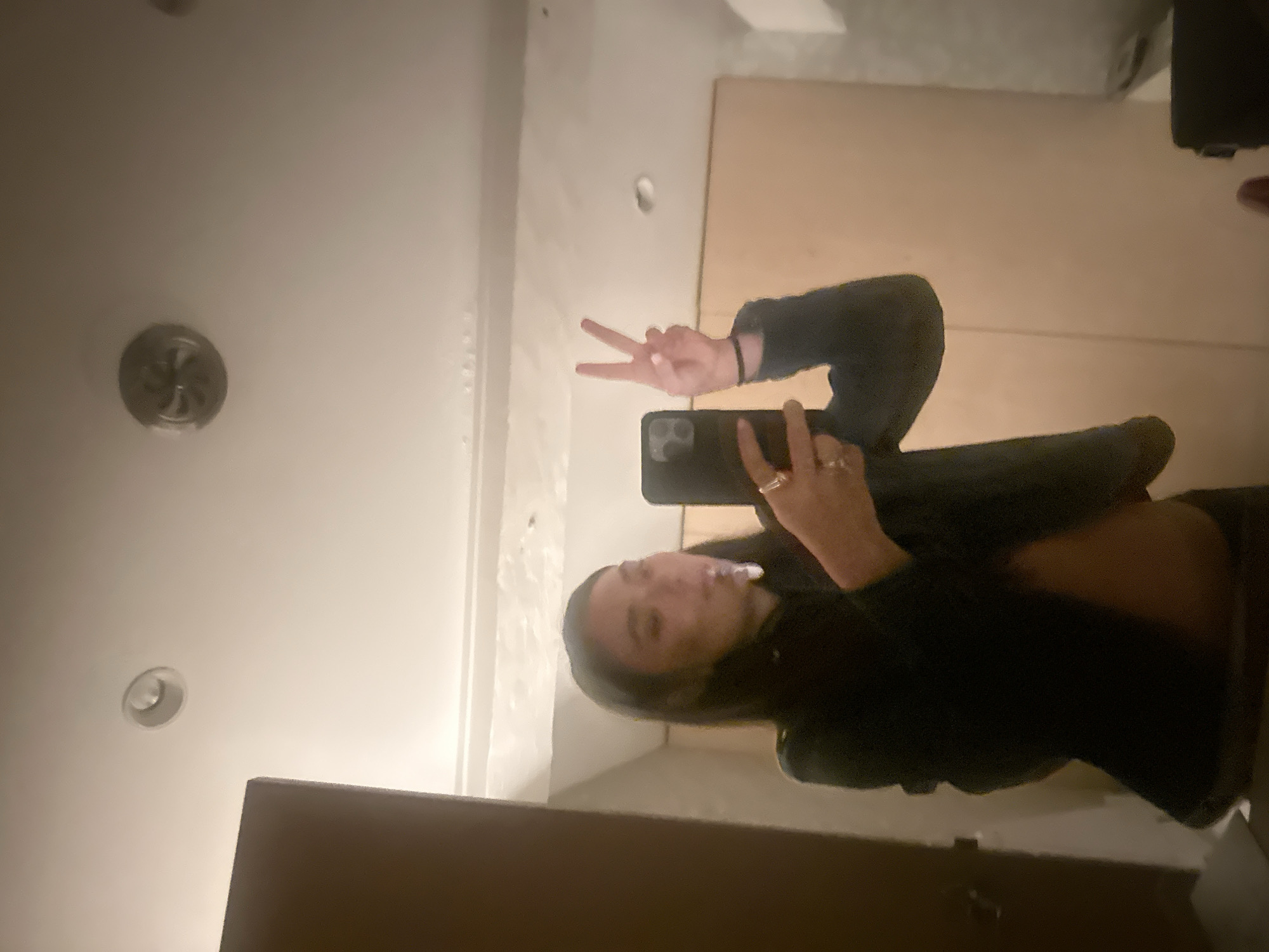 A person stands inside, likely in a bathroom, taking a mirror selfie with a phone. They are making a peace sign. The ceiling has multiple recessed lights. A section of a door is visible. The location is unknown and cannot be confirmed as Paris, France.