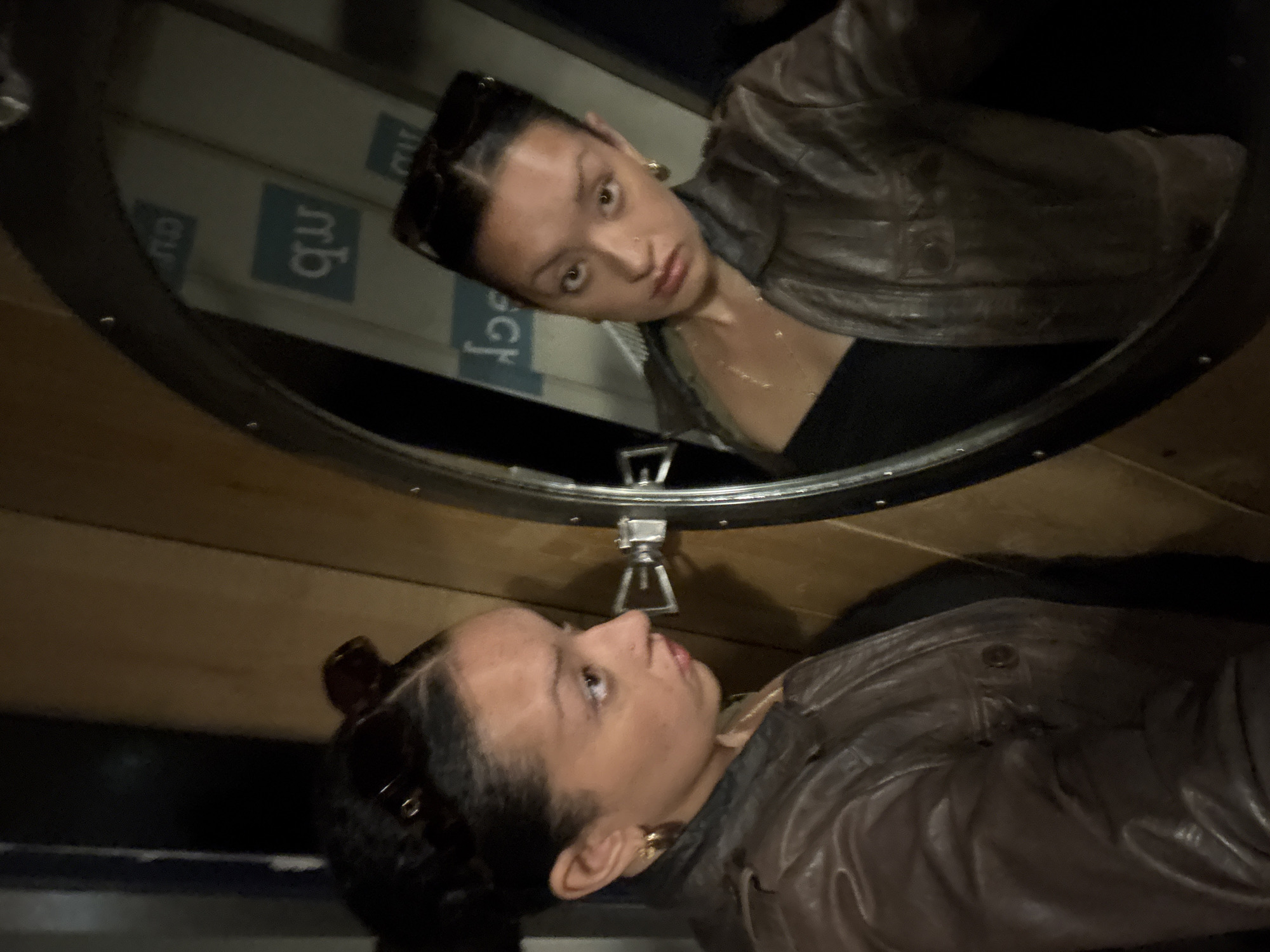 A person and their reflection are captured in a circular mirror in an indoor setting. The person, appearing young, has dark hair tied back with sunglasses resting on top, wears gold hoop earrings, a thin gold necklace, a dark top, and a brown/grey leather-like jacket.
Within the circular mirror, the person's reflection shows their face looking directly towards the viewer with a neutral, slightly serious expression. Outside the mirror, the person is seen in profile, looking upwards and to the right, towards the mirror's surface. The mirror itself has a dark, possibly metallic frame, reminiscent of a porthole, and a prominent metal latch mechanism is visible directly below it.
The immediate surroundings of the mirror consist of light-colored wooden panels. The background reflected in the mirror shows vertical surfaces with visible text. The characters "nb" are clearly legible and upright in the reflection, alongside an inverted "cu" or "eu" to their left, suggesting signage or branding in the environment behind the person.
The scene appears to be dimly lit, possibly at night or in an interior space with controlled artificial lighting. The combination of the porthole-style mirror, wooden paneling, and the specific text cues ("nb", inverted "cu") points to a distinctive interior. Given the location context of London, United Kingdom, this could be a themed establishment like a unique pub, cafe, or potentially an interior of a vessel or structure with a nautical or vintage design.