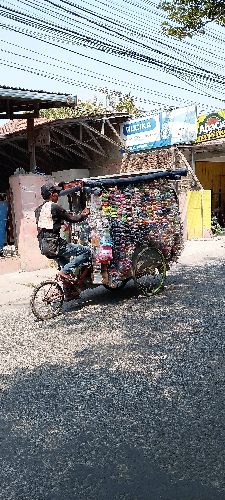 A street vendor is riding a three-wheeled bicycle cart filled with various items, possibly accessories like hair clips and small toys, down a paved street. The vendor, wearing a black long-sleeved shirt, blue jeans, and a black baseball cap, has a white towel draped over their shoulder and a dark bag slung across their chest. The cart itself is a colorful display of merchandise, neatly arranged on racks. In the background, buildings with corrugated metal roofs and brick walls are visible, along with overhead power lines and trees. A sign for "RUCICA" and another partially visible sign for "Abacia SHAWARMA" are also present, suggesting a commercial area. The scene takes place in Babelan, Indonesia, indicated by the presence of signs in Indonesian and the overall context of a street market.