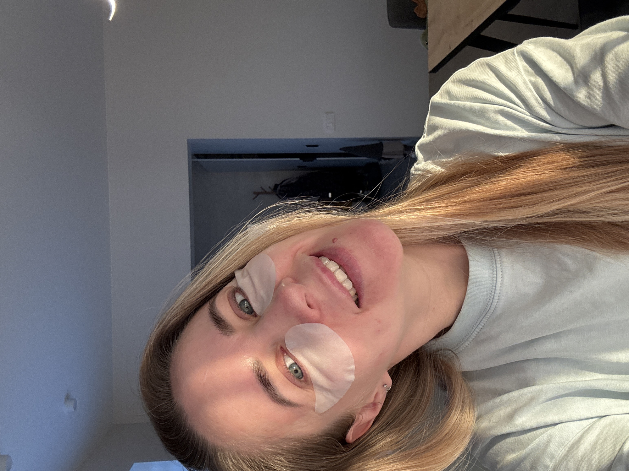 The image features a young woman with long, straight, light-blonde hair, captured in an indoor setting. The photo is rotated ninety degrees counter-clockwise, presenting the woman looking upwards with her head tilted. She is smiling broadly, revealing white teeth, and her eyes are a striking blue-green. Under each eye, she wears a light, translucent, crescent-shaped patch, commonly used for beauty treatments like lash extensions or hydrating eye masks.
She is wearing a light blue, long-sleeved t-shirt with visible stitching detail around the neckline and shoulder. A small silver stud earring is visible in her left ear. Her complexion is clear, with a slight flush on her cheeks, and a small mole is visible above her upper lip. The lighting suggests natural daylight, possibly coming from a window to her left, creating highlights on her hair and face.
The background consists of plain, light-colored walls, appearing off-white or light grey. In the distance, an unidentified dark, rectangular object, possibly a TV screen or a dark piece of furniture, is visible. Above this object, an electrical outlet is faintly discernible on the wall. A bright, crescent-shaped light source or reflection is noticeable in the upper left corner of the frame.
The woman appears happy and relaxed, engaging with the camera. The activity taking place is likely a self-care routine or a beauty treatment involving the eye patches, while she poses for a photo. There is no visible text in the image. Although the context mentions Chaikovsky, Russia, no specific visual cues within the image confirm this location.