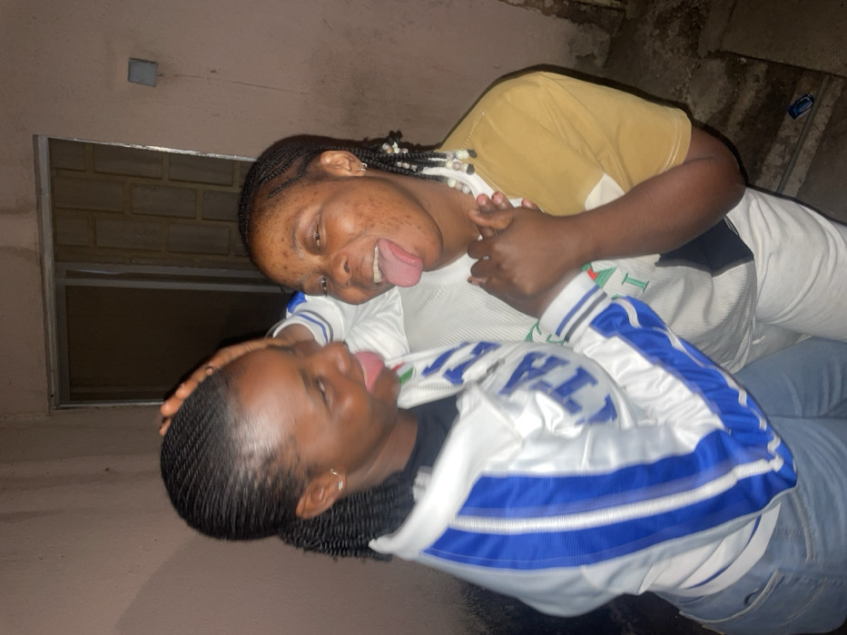 Two young women playfully stick their tongues out toward each other. One woman wears a tan shirt with a white and black-trimmed garment underneath and the other wears a white jacket with blue stripes. Both have dark braided hair. The photo is taken at an angle. A window is visible in the upper left corner and a concrete-like wall forms the backdrop.