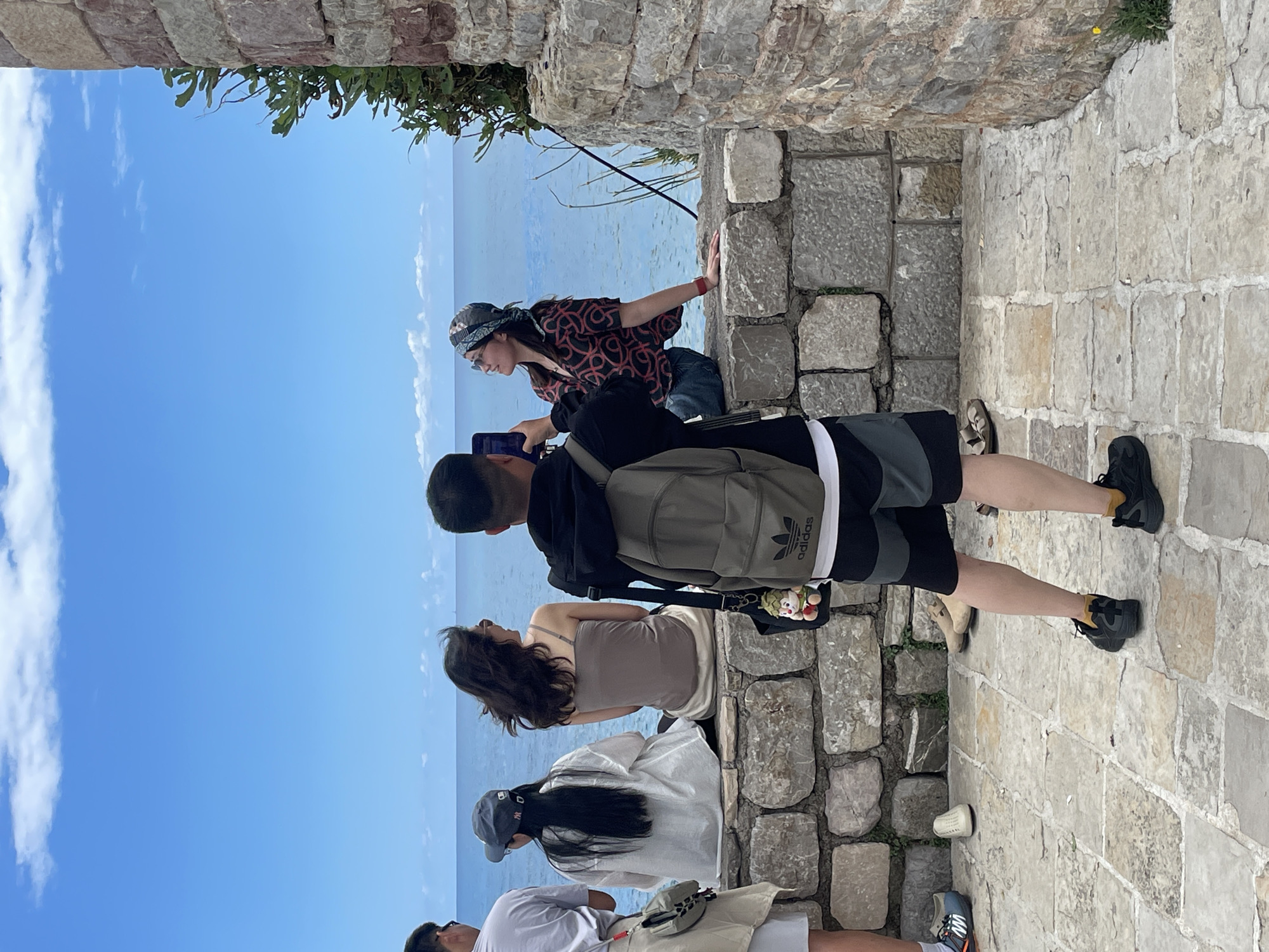 This vibrant daytime image captures a group of people enjoying a scenic coastal view. The scene is set outdoors under a clear blue sky, partially adorned with fluffy white clouds, and a vast, calm blue sea extending to the horizon.
In the foreground, a rugged stone pathway, composed of large, irregularly shaped light-colored blocks, provides the ground for several individuals. To the right, a robust stone wall, built from similar blocks, runs alongside the path, suggesting ancient architecture or a fortified overlook. Overhanging green foliage is visible at the top edge of the image, likely growing from above the stone structure.
Several people are positioned along the pathway, most looking out towards the sea. One person, seen from the back, wears an olive-green Adidas backpack, black shorts with grey and white stripes, and black sneakers with yellow detailing. A small, colorful plush toy or charm hangs from a bag near them. Adjacent to them, another individual with long dark hair wears a beige strapless top. Further left, a person with long dark hair is seen from behind, wearing a white top and a baseball cap. Partially visible on the far left, another individual wears a white shirt and light-colored shorts.
To the right, a person wearing a patterned dark red and black shirt, sunglasses, and a bandana around their head leans against the stone wall, holding a mobile phone in their hand, seemingly capturing the view or looking at content on the device.
The activity suggests sightseeing or observing the natural beauty of the ocean and sky. The bright lighting indicates a sunny day. The only visible text is the "Adidas" logo prominently displayed on the backpack.