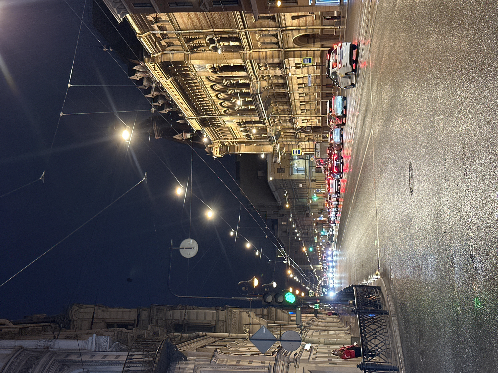 The image depicts a nighttime street scene in Saint Petersburg, Russia, possibly on Nevsky Prospekt, characterized by wet asphalt reflecting streetlights and car headlights. A row of ornate, older buildings with illuminated windows lines the right side of the street, their architectural details visible under the artificial light. On the left, a similar facade of a building with balconies and detailed stonework is partially visible. The street is moderately busy with traffic, showing a line of cars, including a white sedan in the foreground with its brake lights illuminated. A traffic light is visible, displaying a green signal. A woman in a red jacket stands near a black metal fence on the sidewalk to the left. Overhead, a network of wires and streetlights crisscrosses the dark night sky, contributing to the urban atmosphere. The overall impression is of a vibrant, wet city at night.
