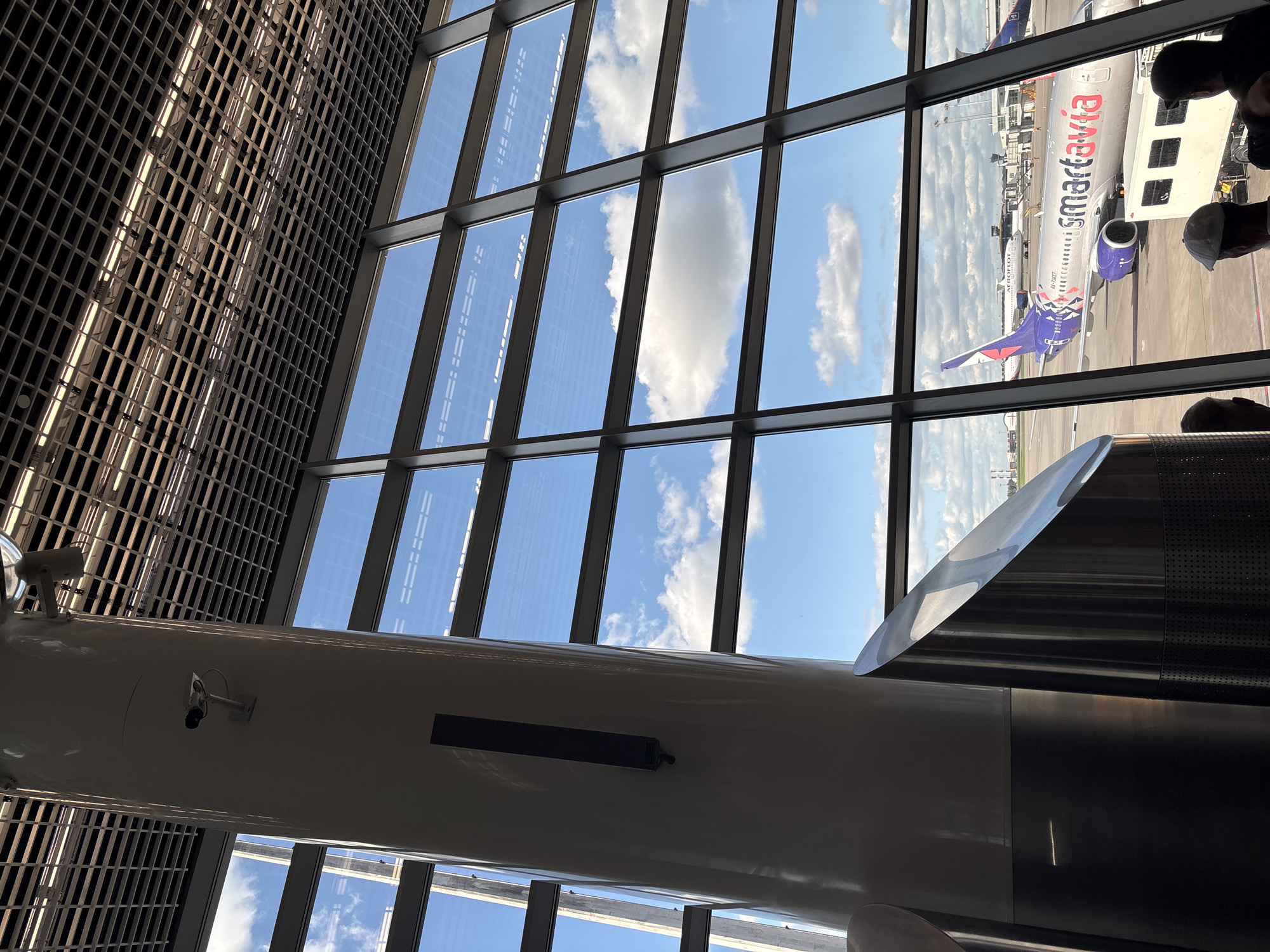 The image depicts an airport scene viewed through a large window comprised of multiple panes. The window's metal frame creates a grid-like pattern against a bright blue sky with scattered white clouds. Outside the window, an airplane with the "smartavia" logo is visible on the tarmac. There are passengers near the plane. The building has interior design, including cylindrical supporting columns with mounted fixtures like security cameras. The overall composition suggests the interior of a modern airport terminal.