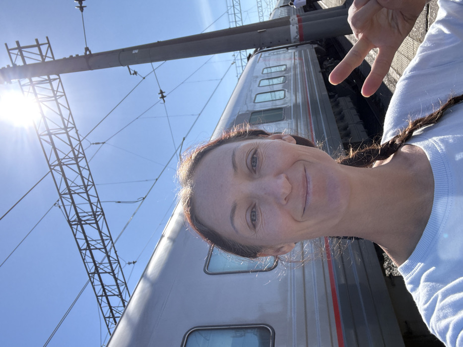 A forensic description of the scene depicts a single adult female in the foreground, centered and looking directly at the viewer with a slight smile. She has light skin, dark hair styled in a braid over her right shoulder, and is wearing a light-colored, long-sleeved garment. Her left hand is raised, displaying a "peace" sign (V-sign) with her index and middle fingers extended.
The background features a silver-grey passenger train with multiple rectangular windows, running horizontally across the middle ground. A red stripe is visible along the lower portion of the train's body. Overhead, a clear blue sky dominates, with a bright sunburst emanating from the upper right, casting a strong light. An extensive railway electrification system, consisting of dark metal trusses, wires, and a tall vertical pole, is visible against the sky and behind the train. In the far distance to the right, beyond the train and the pole, faint figures of other individuals are discernible on what appears to be a railway platform. The location is a train station or railway stop in Kropachyovo, Russia.