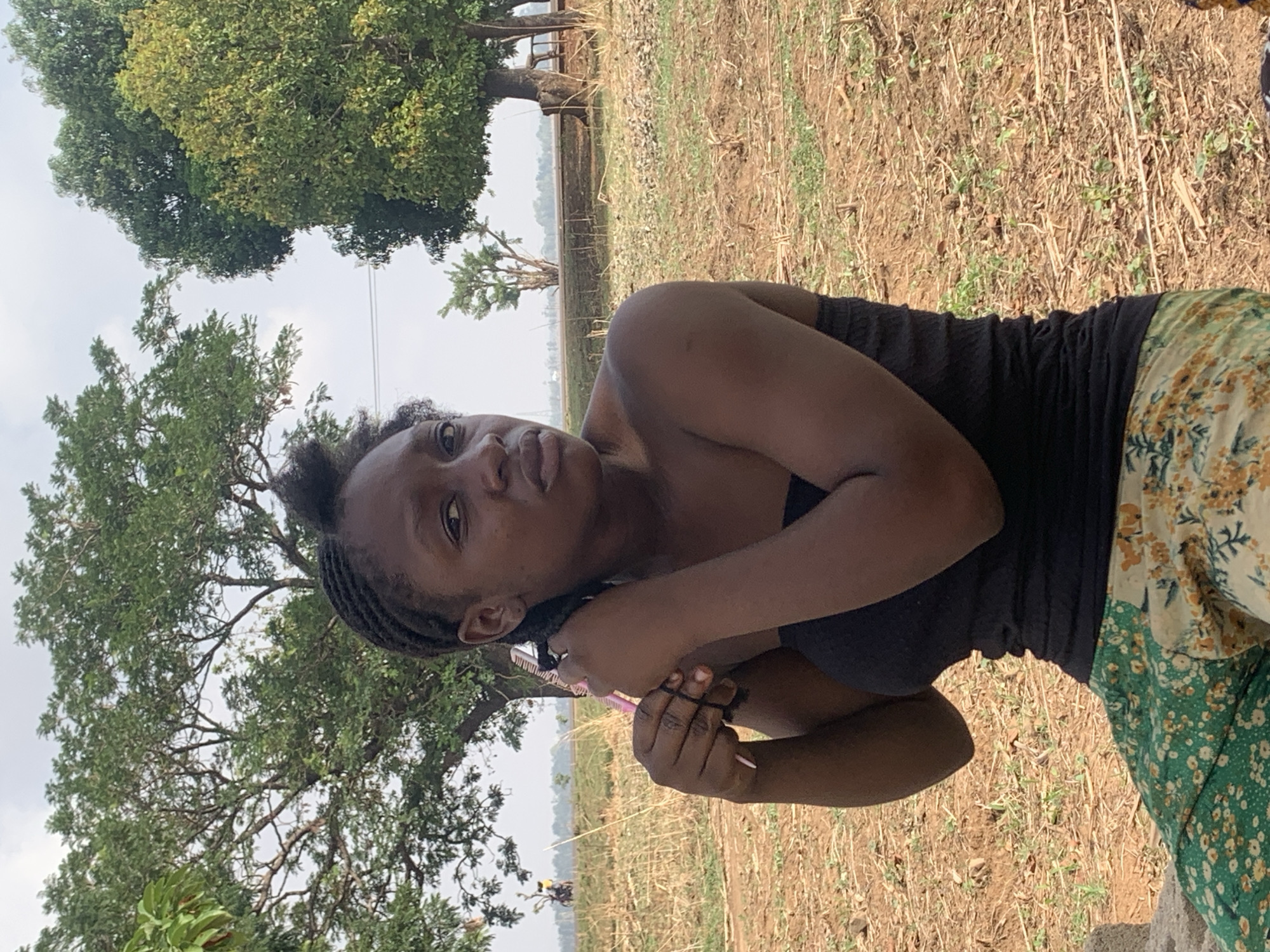 An outdoor, vertically oriented, mid-shot photograph features a young woman of African descent posing in a rural setting in Agban, Nigeria. She stands slightly off-center to the right, looking directly at the viewer with a neutral, calm expression. Her dark hair is styled in braids, and she wears a dark, possibly strapless, top and a flowing skirt adorned with green, yellow, and white floral patterns. Her arms are crossed at chest level, and she holds a slender, light-colored stick or pen in her hands. A small mark is visible on her right shoulder.
The background depicts a dry, earthy field with sparse patches of green vegetation and dry stalks, suggesting an agricultural area or uncultivated land. Several large trees with dense green foliage frame the scene against a light, hazy sky, indicating daytime, likely late morning or early afternoon. In the distant mid-ground, a faint, flat landscape can be seen. The overall atmosphere is natural and calm, capturing a moment in a sun-drenched, rural environment. No text is visible in the image.