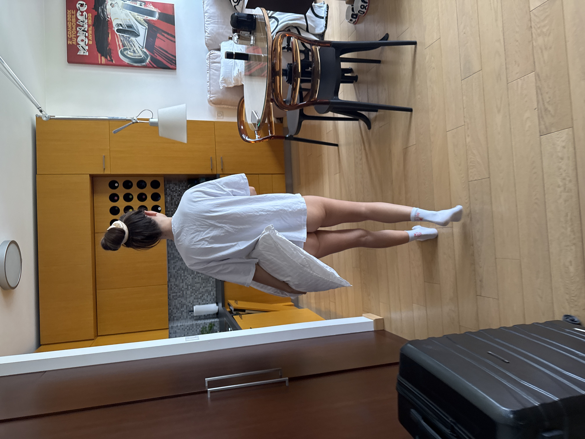 A rear view of a woman is captured walking across a light-colored wooden plank floor towards a kitchen and dining area in a modern apartment, located in the city of Unknown, Unknown. The woman has her dark hair tied in a high bun with a light-colored scrunchie. She is wearing an oversized white t-shirt that extends to mid-thigh, revealing bare skin beneath, and white ankle socks with faint red lettering on them. She carries a white pillow under her left arm.
To the left, a dark brown full-height cabinet or doorway with two horizontal silver handles is partially visible. The kitchen area features flat-panel mustard yellow cabinetry with silver bar pulls, a dark countertop, and a grey tiled backsplash. An integrated wine rack with dark bottles is visible within the upper cabinets. A roll of white paper towels stands on the counter.
In the mid-ground to the right, a round glass-top table with a dark pedestal base is surrounded by two modern chairs. One chair has a dark frame with a translucent smoky brown back and seat; the other is mostly dark. On the table, a dark cylindrical container, a clear glass bottle, and a smartphone are placed. Behind the table, a cream-colored sofa is partially visible, with a white throw blanket patterned with black shapes draped over its arm. A pair of white slippers with black lettering is on the floor near the sofa.
A large, rectangular red poster depicting a vintage silver race car and text that reads "2EME GRAND PRIX AUTOMOBILE MONACO 5 AVRIL 1930" hangs on the white wall above the sofa. An articulated ceiling lamp with a conical white lampshade extends from the ceiling over the dining table area. The ceiling is white, featuring a round, flush-mount light fixture. A black hard-shell rolling suitcase is partially visible in the bottom-left foreground. The apartment is well-lit, suggesting natural light from an unseen source.