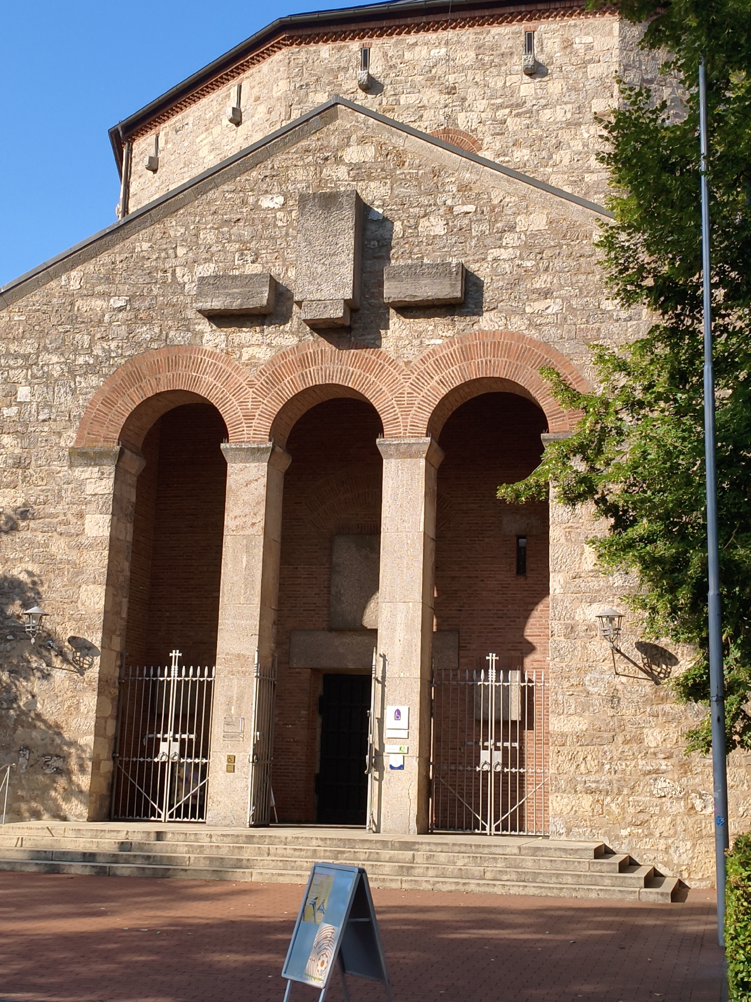 The image shows the facade of a stone church, possibly in Bamberg, Germany. The church features three large arched entrances supported by thick stone columns. The entrances are partially obscured by ornate metal gates. The stonework of the church is rough-hewn, with smaller stones used for the arches. A large tree with green foliage is visible on the right side of the image, partially obscuring the church. In front of the church, there are stone steps leading up to the entrances, and a paved area with a sidewalk. An A-frame sign with graphics is positioned in the foreground, slightly to the right of the center. The sky is a clear, bright blue. The overall scene suggests a sunny day.