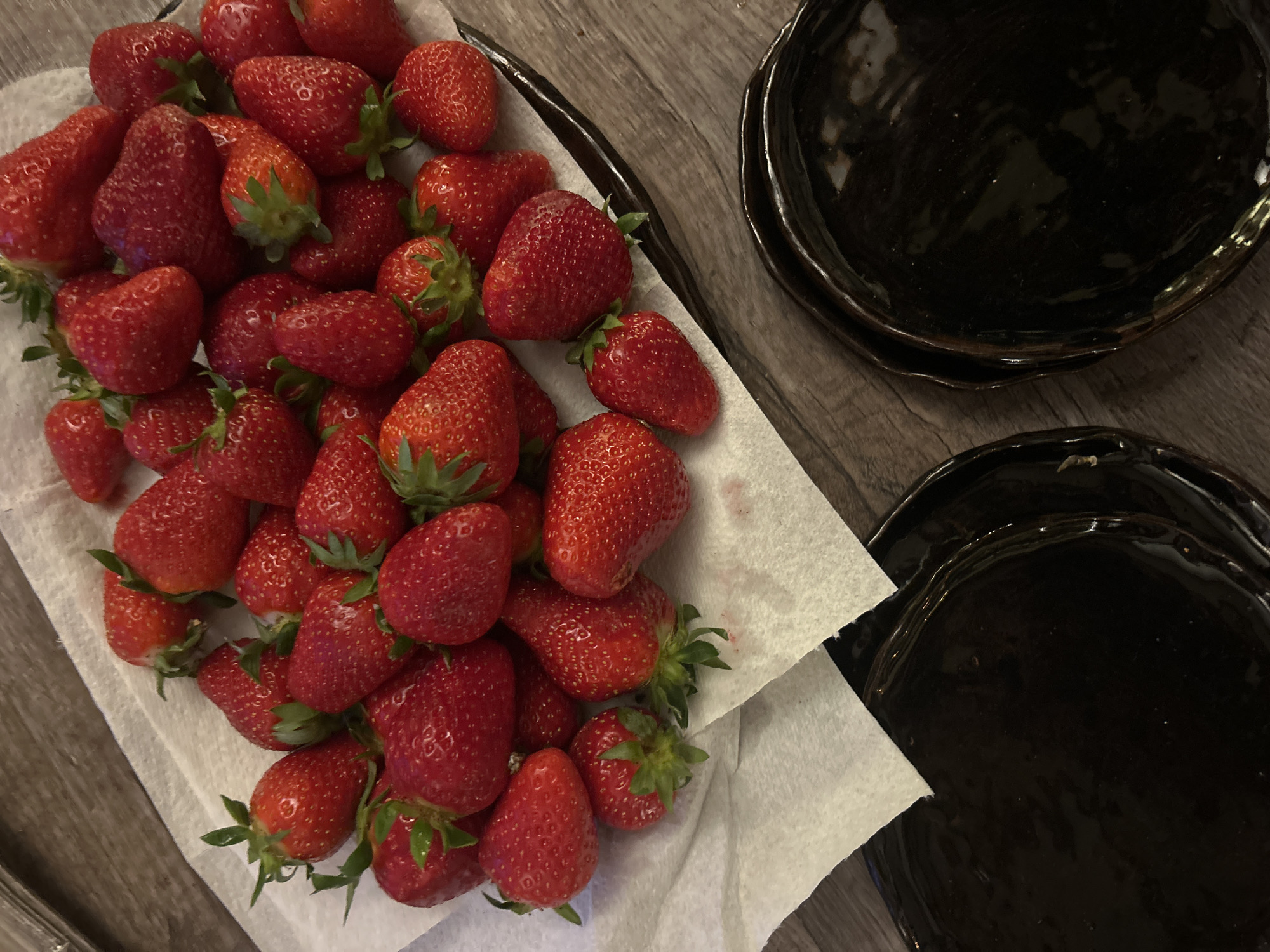 A high-angle shot shows a collection of fresh strawberries presented on a square of white paper towels within a dark gray or black rimmed serving platter. To the right of the strawberries are two dark, glossy plates stacked together, with another single plate below them. All items are placed on a surface that appears to be wood-like with a gray or light brown color. There is no clear indication of the location or any individuals.