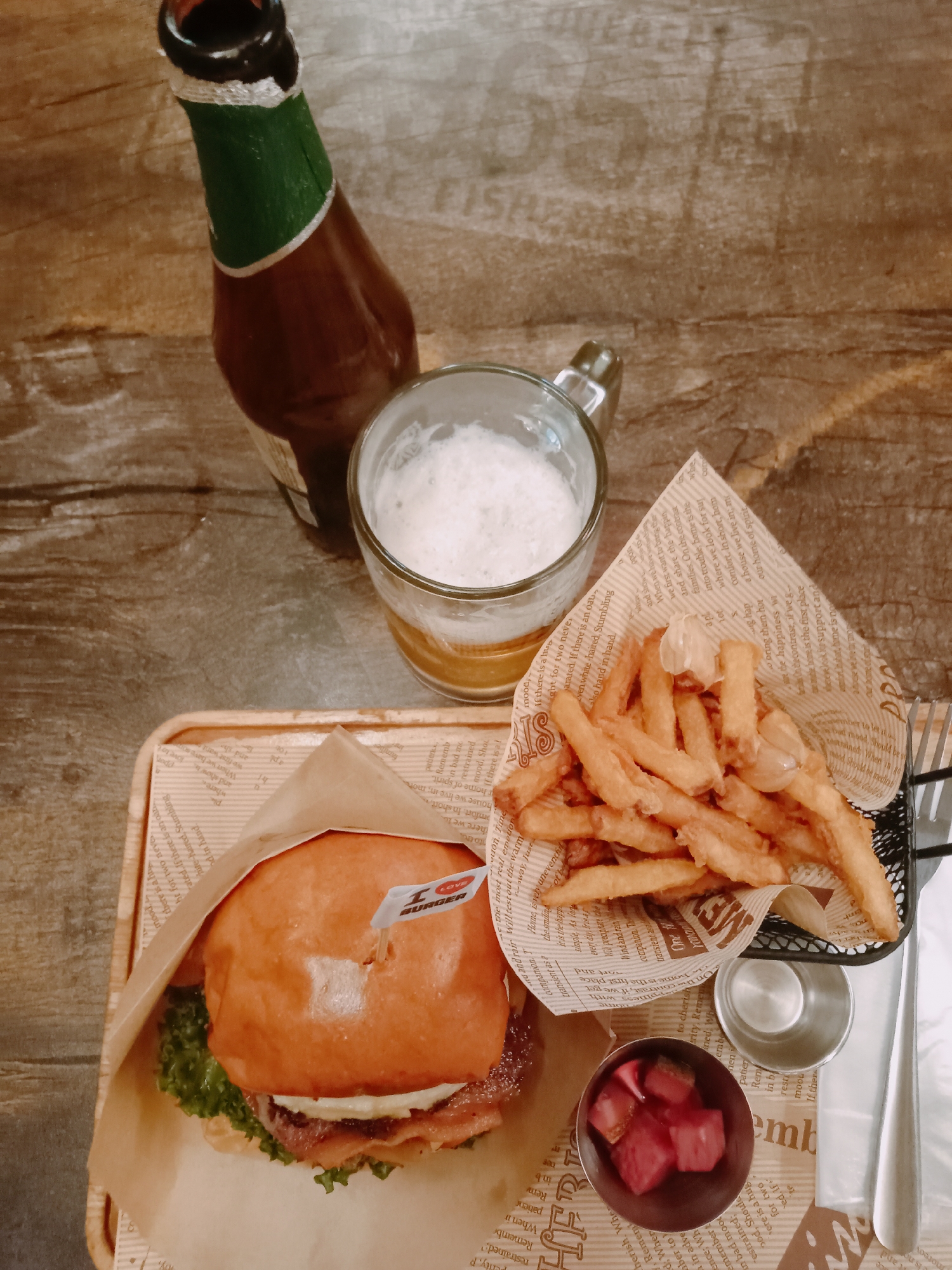 Captured from an elevated, perpendicular vantage point, the scene presents a culinary ensemble arranged on what appears to be a textured wooden surface, possibly a table in Nha Trang Ward, Vietnam. The composition features a burger with an "I LOVE BURGER" flag embedded, alongside golden-brown French fries presented in a newspaper-lined container, a small bowl of pickled items, and silverware. Complementing the meal are a glass mug filled with beer and a dark-colored beer bottle. The arrangement suggests a casual dining setting.