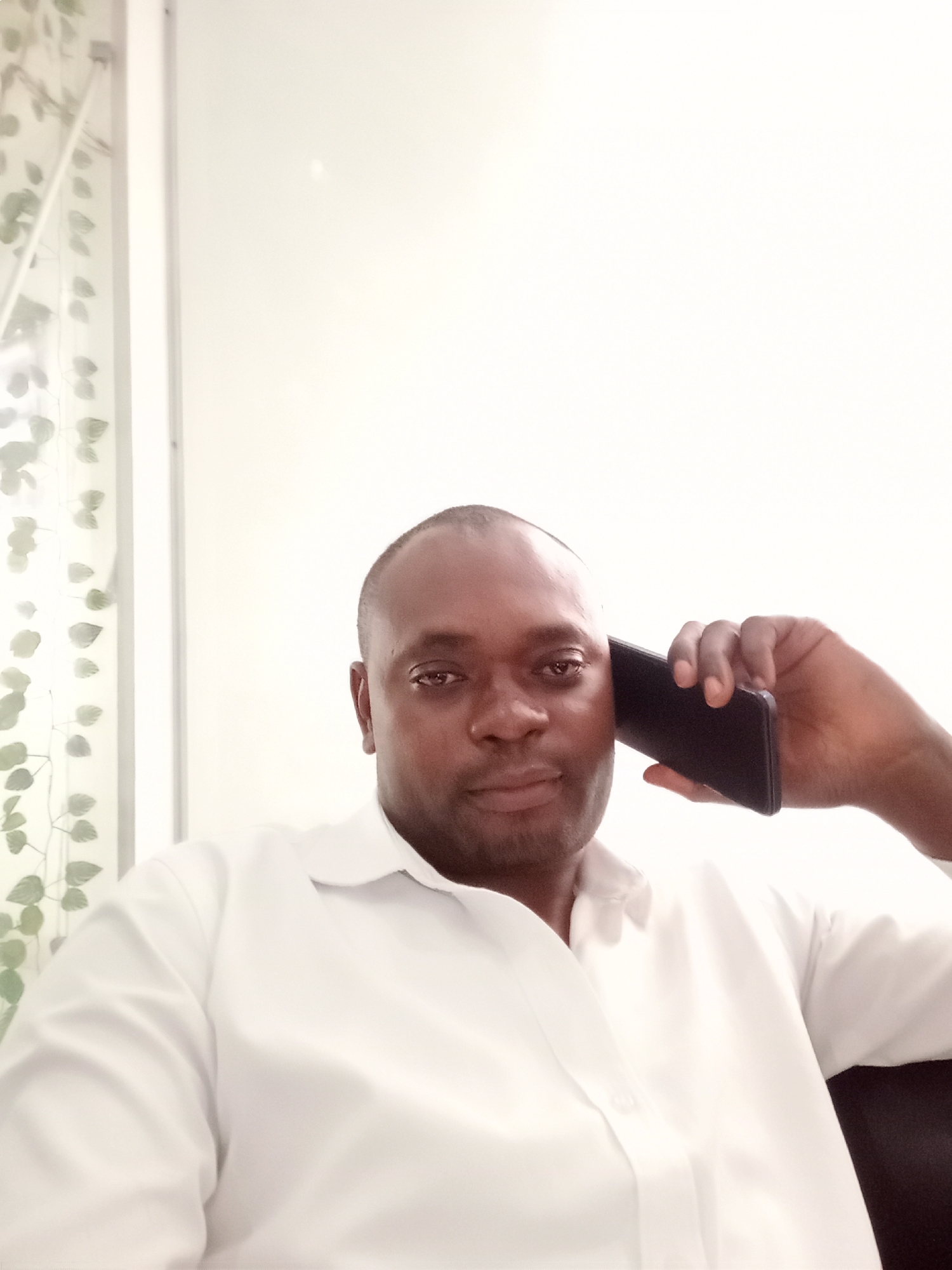 An adult man is featured in a selfie-style portrait, holding a black smartphone to his right ear with his right hand, seemingly engaged in a phone call. He has a shaved or very short haircut and is wearing a crisp white long-sleeved collared shirt, with the top button unfastened. His expression is neutral, with a slight hint of a smile, as he looks directly at the camera.
The background to his left shows a narrow vertical white or light-colored frame, possibly part of a window or door, beyond which blurred green and grey leaves, resembling decorative artificial vines, are visible against a bright backdrop. The rest of the background, primarily to his right and above, is a plain, brightly lit white wall, which appears slightly overexposed due to the strong illumination. The overall setting appears to be indoors. No text is visible in the image. The context for the location is Gwarinpa, Nigeria.