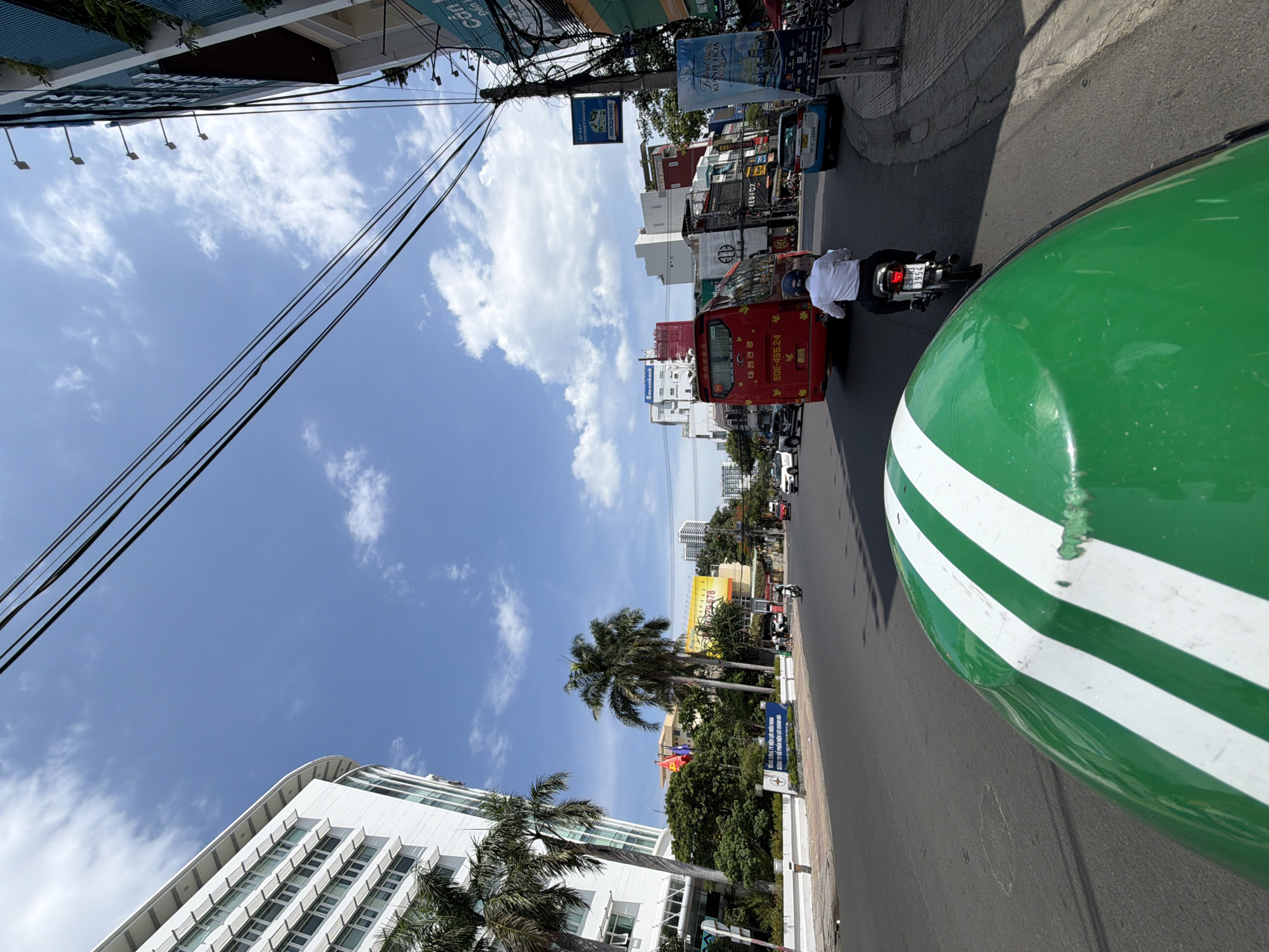 The image captures a street scene in Nha Trang, Vietnam, on a sunny day with scattered clouds. In the foreground, the curved, green surface with white stripes of a vehicle's hood dominates the lower portion of the frame. Beyond this, a two-lane asphalt road stretches into the distance.
On the right side of the road, a pedestrian sidewalk is visible, lined with buildings featuring signage and advertisements. A man on a white motorcycle, wearing a white shirt and dark pants, is heading away from the camera in the right lane. Behind him, a red bus with "70" prominently displayed on its rear is traveling in the same direction. Several cars and other vehicles are also present on the road.
To the left of the road, a large, modern white building with many windows is a prominent feature, with several palm trees in front of it. Further down the street, more buildings and trees are visible, indicative of an urban environment. Power lines crisscross the sky, a common sight in Vietnamese cities. The overall atmosphere suggests a bustling city street during daylight hours.