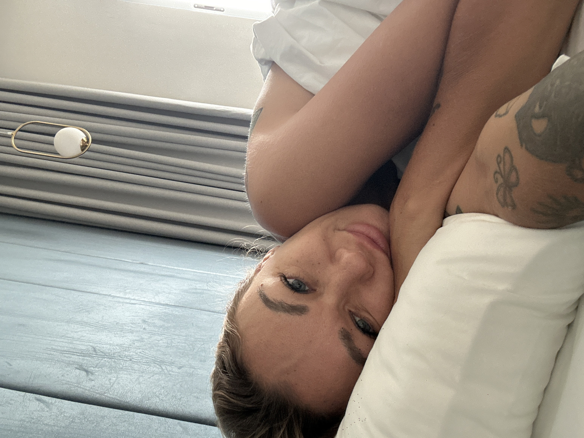 A person lies on a white pillow. Their face is tilted towards the viewer, and their expression is neutral. The person has blonde hair and blue eyes. Their arms are crossed, and tattoos are visible on their left arm. A wall lamp with a white globe and gold frame is in the background, along with gray drapes and a light blue wooden floor. The location information is not available in the image.