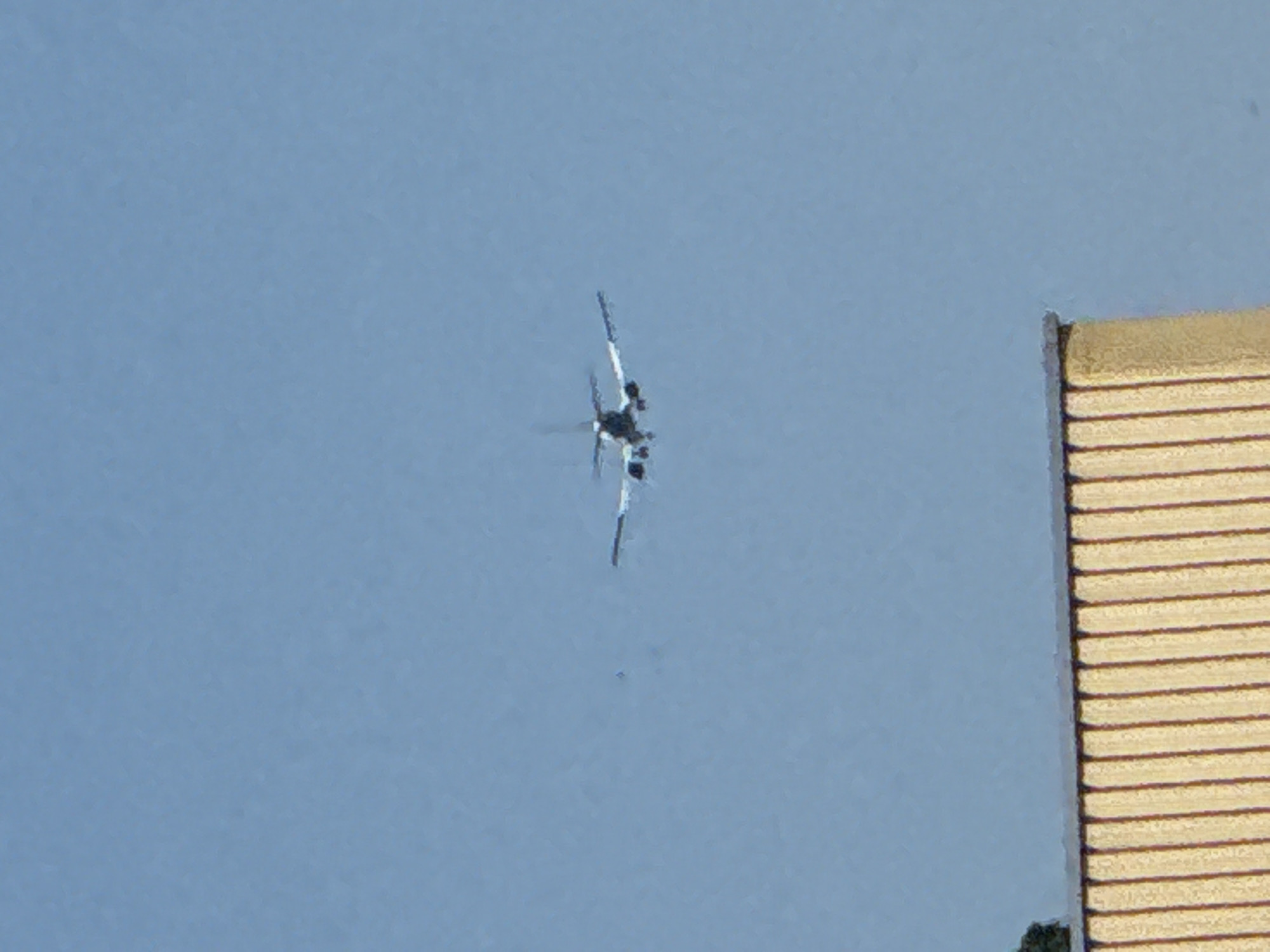 An airplane is visible in the sky, slightly off-center to the right. It is in flight with its landing gear extended. In the foreground, the top of a building with vertical, light-colored panels and a dark roofline is present. The background is a clear blue sky. There is no discernible location information within the image itself.