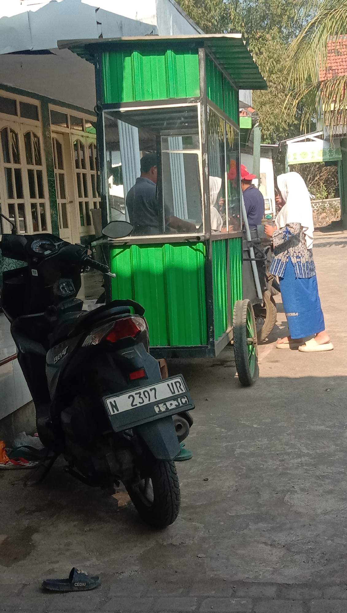 In Sidogiri, Indonesia, a black motorcycle is parked on a concrete surface in front of a green food stall. The stall, constructed with metal and green corrugated metal siding, features a glass enclosure. Inside the stall, a man wearing a dark shirt is visible, seemingly serving customers. Two women stand near the stall. One woman, wearing a white hijab and a patterned top and blue skirt, is facing away from the camera. Another woman, partially obscured, wears a blue shirt and a red cap. To the left of the stall, a building with white doors and windows is visible. The scene appears to be outdoors during daylight hours.