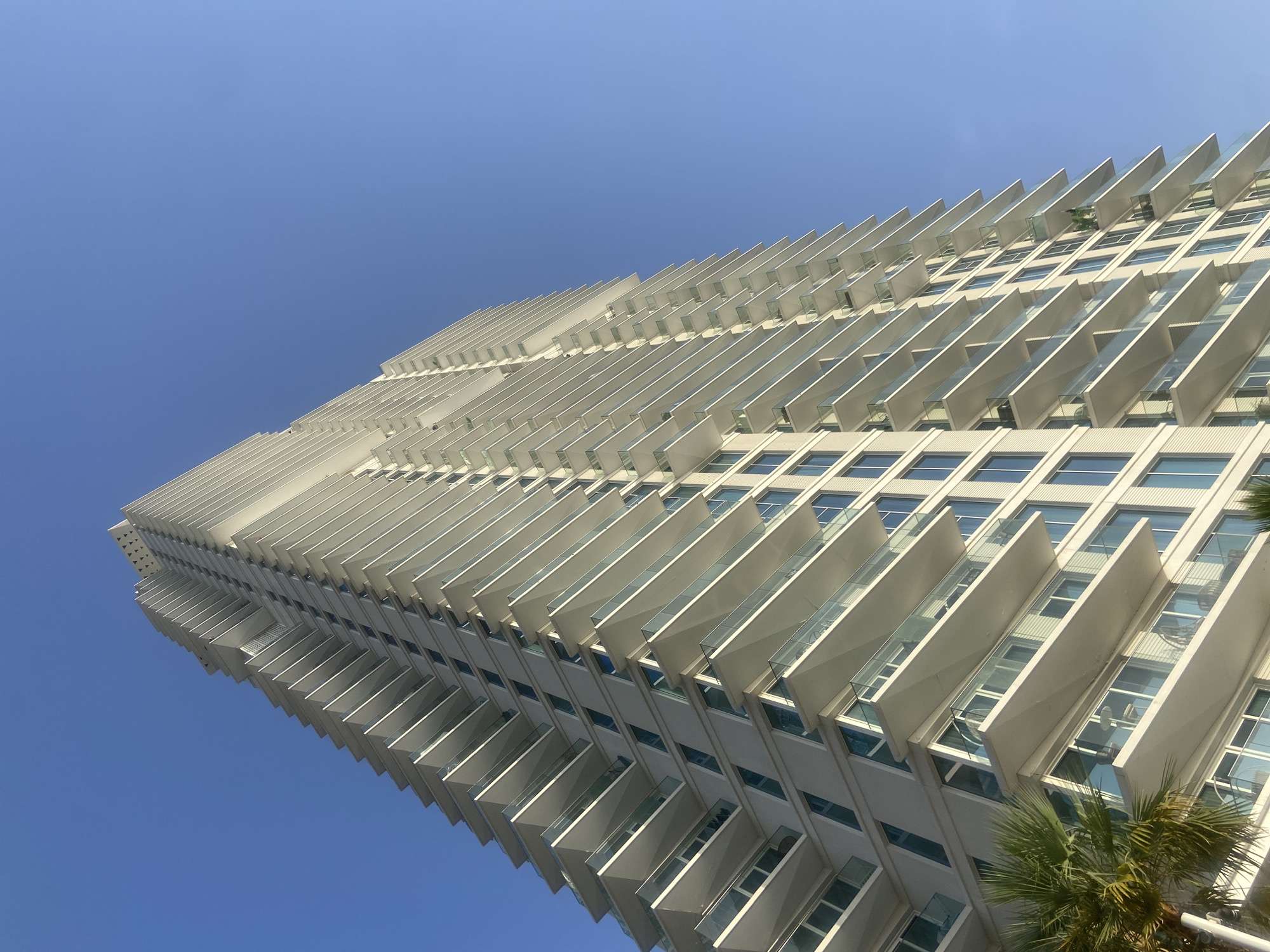 A low-angle view captures a tall, modern building against a clear blue sky, likely in Dubai, United Arab Emirates. The building's facade is characterized by rows of projecting, rectangular structures, likely balconies, that create a staggered, layered appearance. Windows are visible between the projections, and the building's color scheme is predominantly white or light gray. The architectural style is contemporary and angular. A palm tree is visible in the lower right corner.