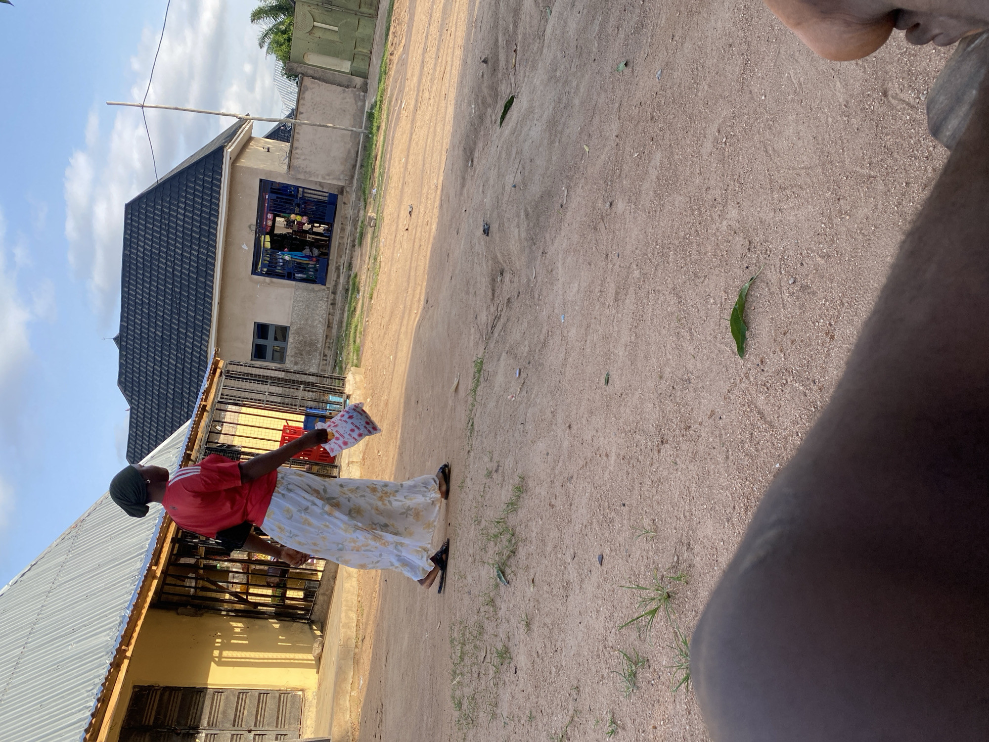 In Howayi, Nigeria, a woman walks on a dusty dirt road. She wears a red t-shirt with white stripes on the sleeves, a headscarf, and a long, floral print skirt. She carries a small, white package in her right hand. Behind her are two buildings: one yellow with a corrugated metal roof and barred windows, and another tan with a dark grey tiled roof and a blue gate. The ground is covered in loose dirt and scattered green grass. The sky is partly cloudy. The foreground of the image is dominated by the legs and feet of an individual, out of focus.