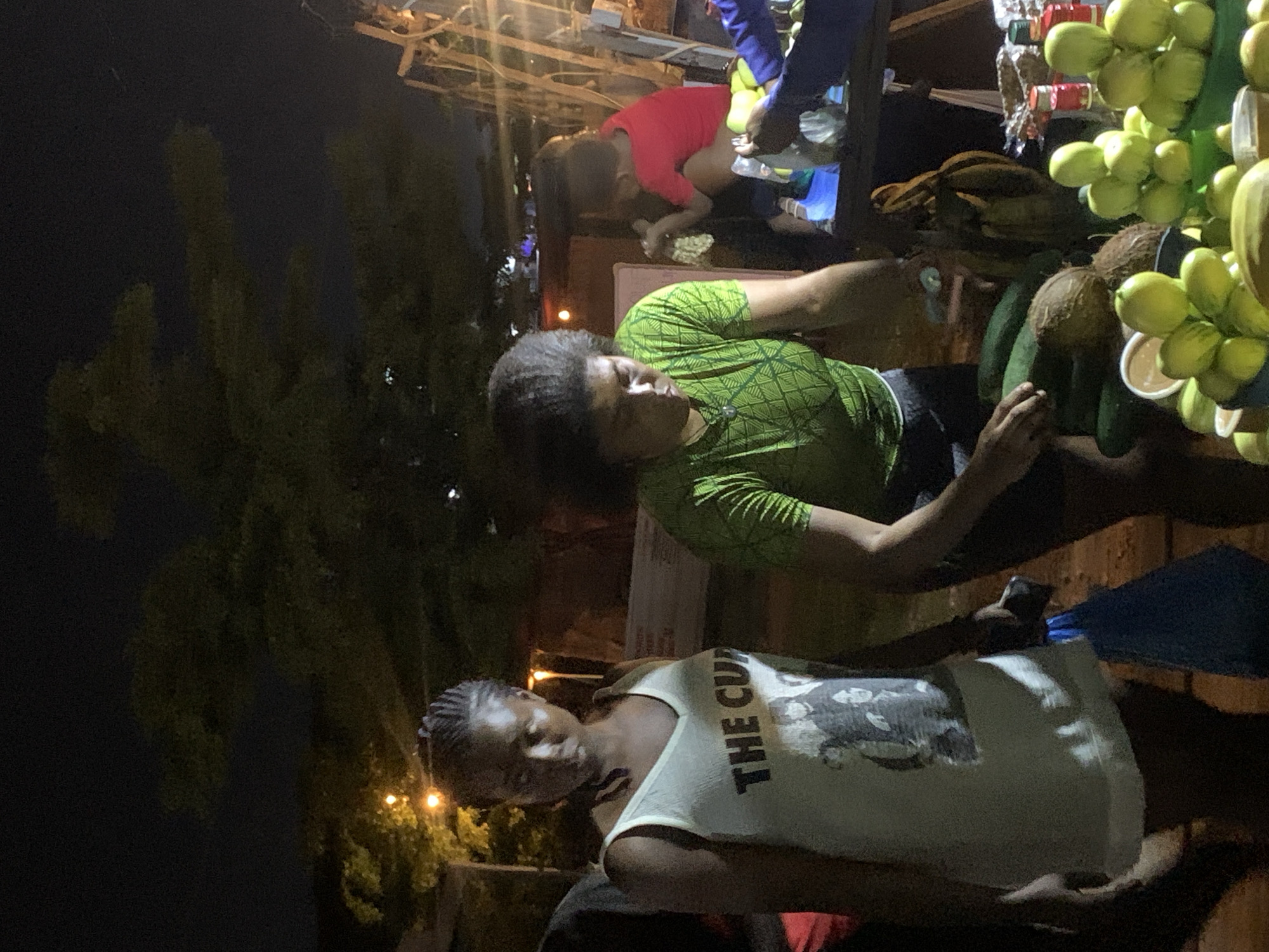In Galadimawa Village, Nigeria, at night, two women are engaged in commerce. The woman on the left, wearing a white tank top with the text "THE CUR" and an image, stands facing forward. To her right, another woman in a green patterned t-shirt bends over a table laden with produce, including coconuts and green vegetables. A child in a red shirt is visible behind them. The background shows trees and a ladder-like structure. The scene appears to be an outdoor market stall, illuminated by artificial light.