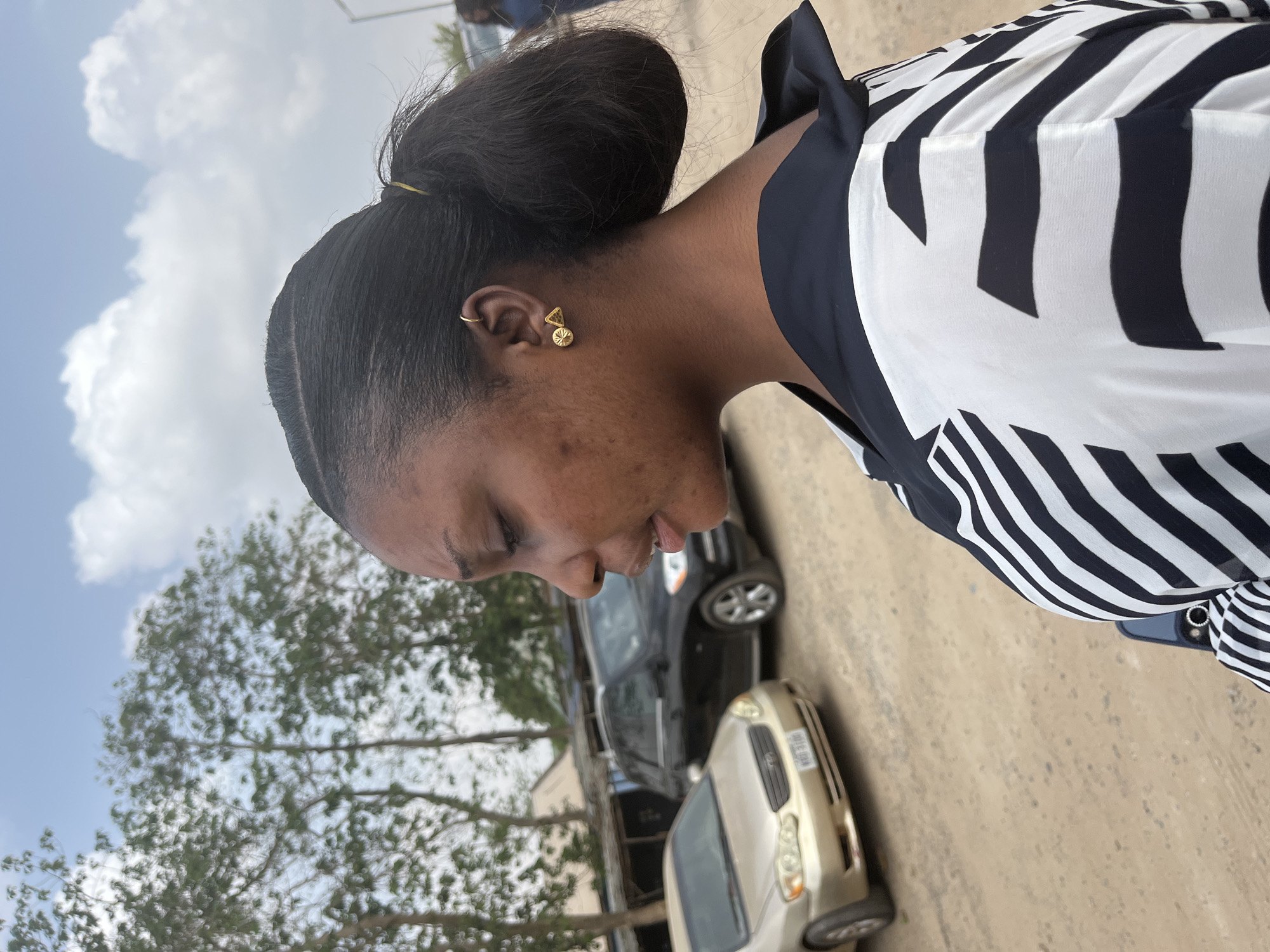 A woman with dark hair pulled back in a bun stands outdoors. She wears a black and white striped blouse and gold earrings. The background includes trees, two cars, and a partly cloudy sky. The location is an urban area of Abuja, Nigeria.