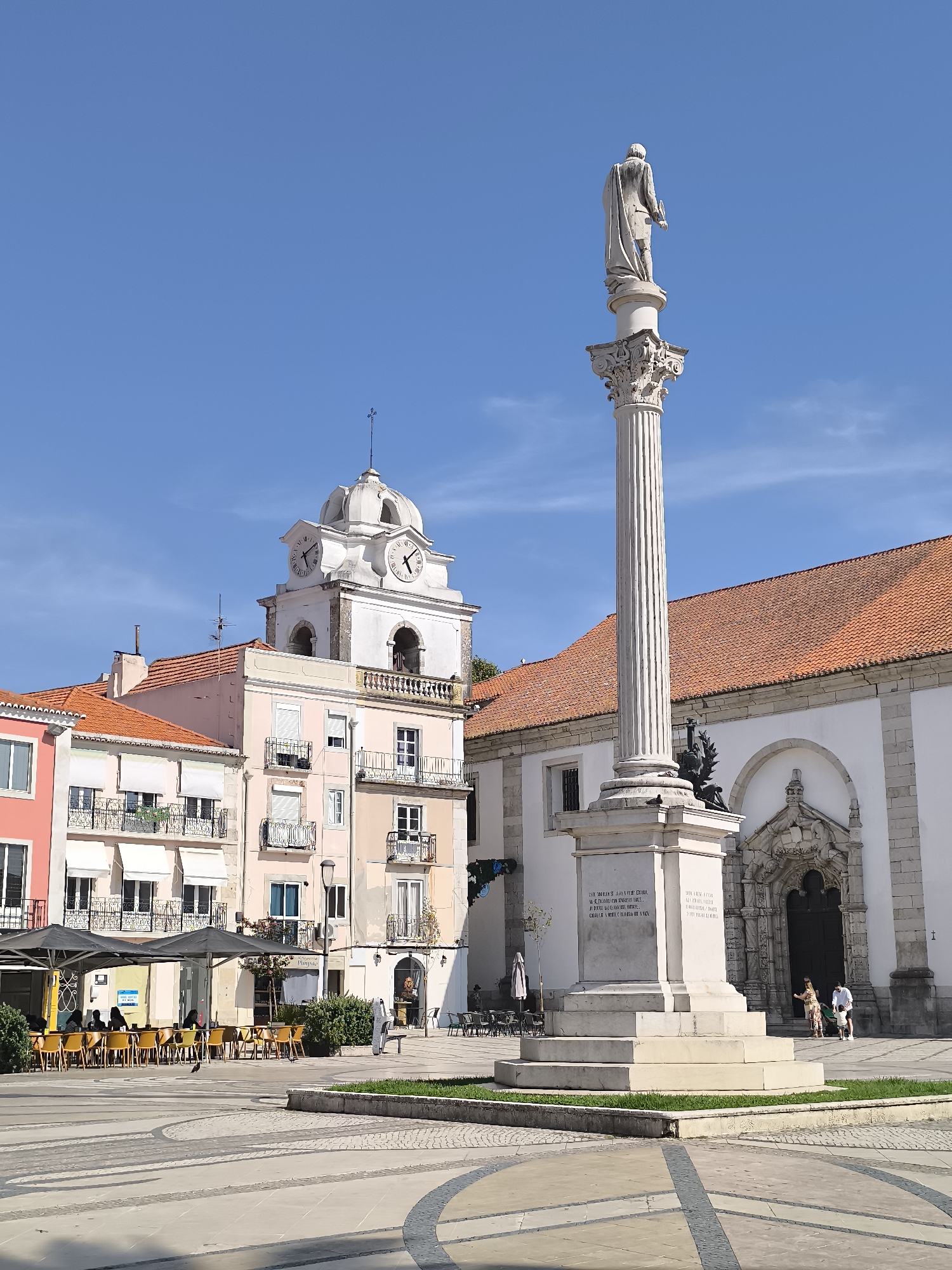 This is a wide shot of Praça de Bocage in Setúbal, Portugal, on a clear day with a bright blue sky. In the foreground, a tall, white marble monument with a statue of a man in a toga standing on top dominates the center of the plaza. The monument has a fluted column and an ornate base with inscriptions. To the left of the monument is a building with a pink facade, balconies with white awnings, and windows. Behind it, a light pink building features a clock tower with Roman numerals and a dome. To the right of the monument, a white building with a red-tiled roof and an arched entrance is visible. Several people are scattered throughout the scene, with some seated at tables outside a cafe under umbrellas, others walking near the church entrance, and a lone figure standing near the monument. The plaza is paved with patterned stone, and there are patches of green grass around the base of the monument.