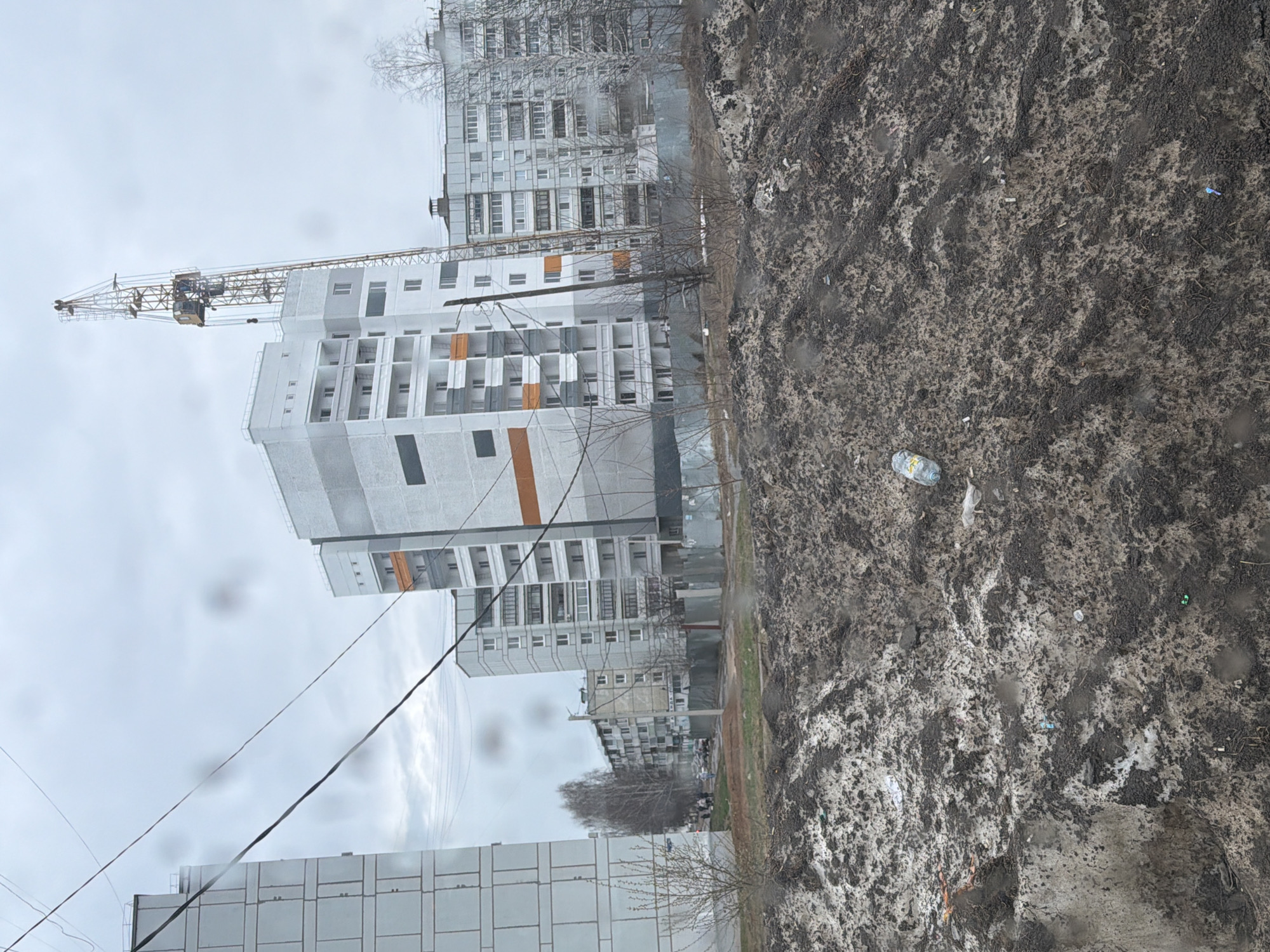 The image depicts a cityscape in Chaikovsky, Russia, under an overcast sky. Several multi-story apartment buildings, constructed primarily of concrete, stand in the background. A yellow construction crane is visible near one of the newer buildings. In the foreground, a dirty, muddy patch of ground is littered with trash, including plastic bottles and other debris. Power lines are strung between the buildings. There are visible droplets on the window from which the photo was taken.