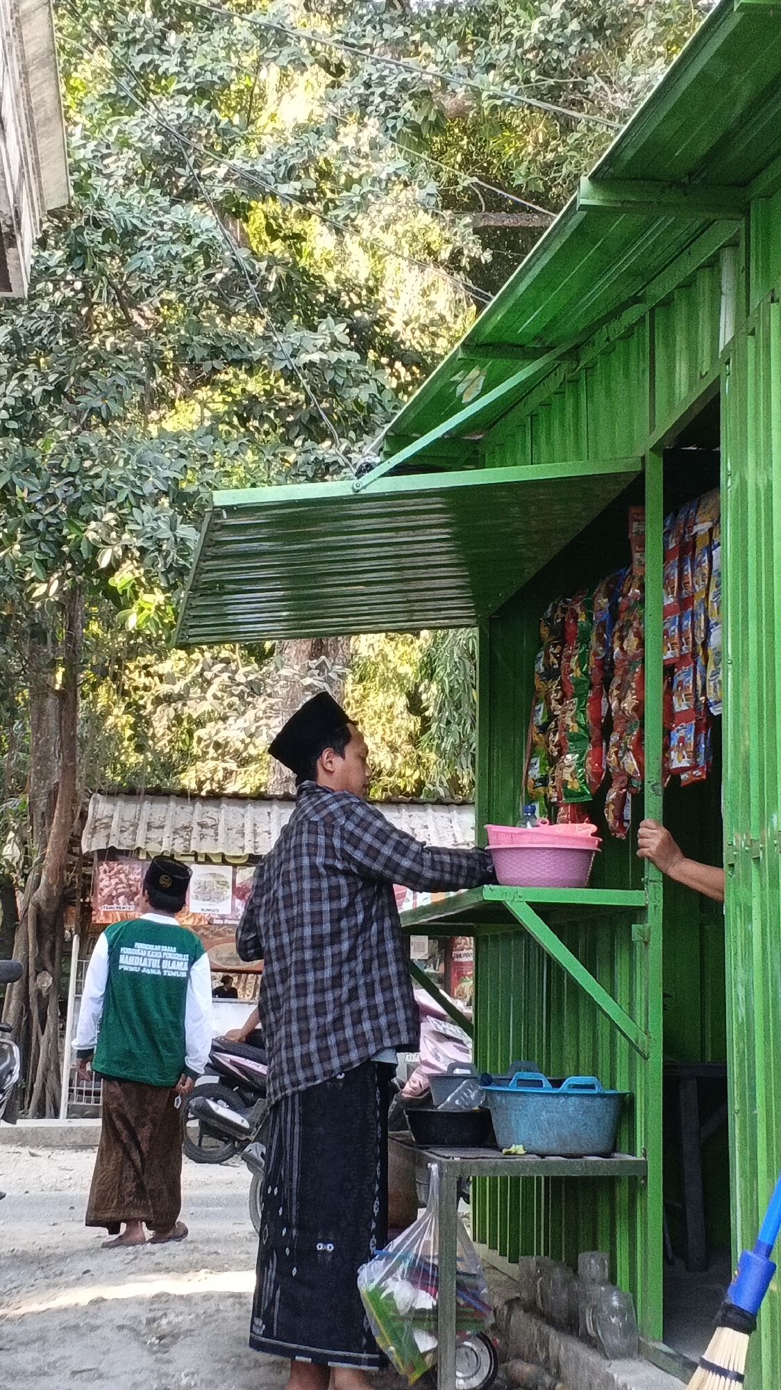 The image depicts a street scene in the city of Rengel, Indonesia. In the foreground, a man wearing a plaid shirt and a black peci (cap) stands at a green stall. He is reaching out to a pink basket on a shelf. Behind him, another man in a green vest and brown sarong also wears a peci. The stall displays various packaged goods on shelves. A motorcycle is parked to the left of the frame. The environment appears to be outdoors with trees and sunlight filtering through the leaves.