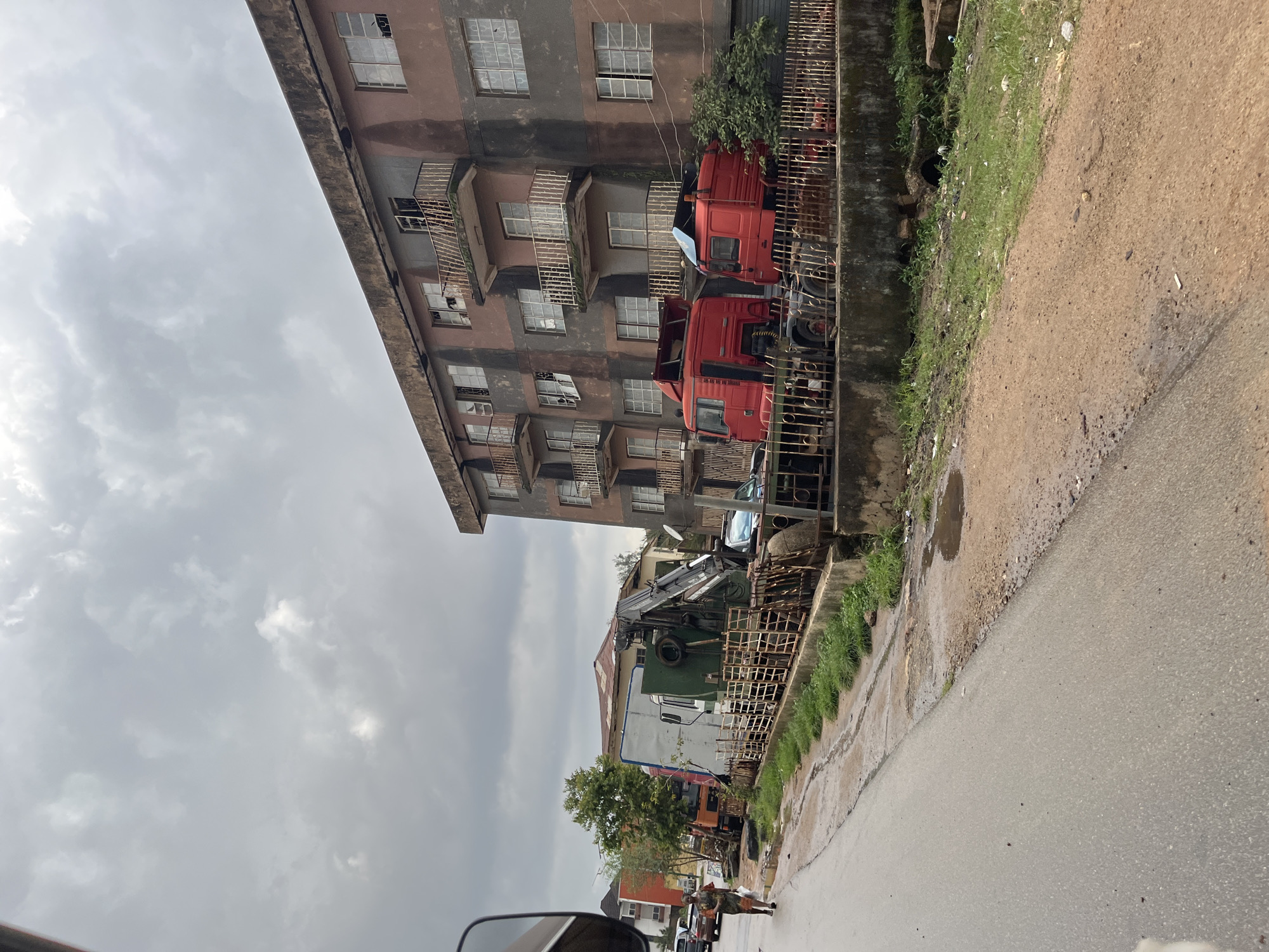 The image depicts a street scene in Jos, Nigeria, under an overcast sky, suggesting a cloudy or potentially rainy day. The perspective is from a vehicle, looking down a wet, paved road. To the left of the road, a multi-story building with a weathered, brownish facade stands. The building has numerous windows, many of which appear to be broken or boarded up. Balconies with rusted railings are visible on several floors.
Adjacent to the building and separated by a metal fence, several large red trucks are parked. One truck is positioned behind another, its cab clearly visible. Further down the street, a larger vehicle with a crane is parked, possibly a construction or utility vehicle. The area appears to be a mix of residential and commercial, with glimpses of other buildings and trees in the background.
The road surface shows signs of recent rain, with puddles and wet patches. The ground alongside the road is covered in patchy green grass and some debris. A person is walking on the right side of the road, wearing an orange top and dark trousers. The overall impression is one of a somewhat neglected urban environment. No visible text is present in the image.