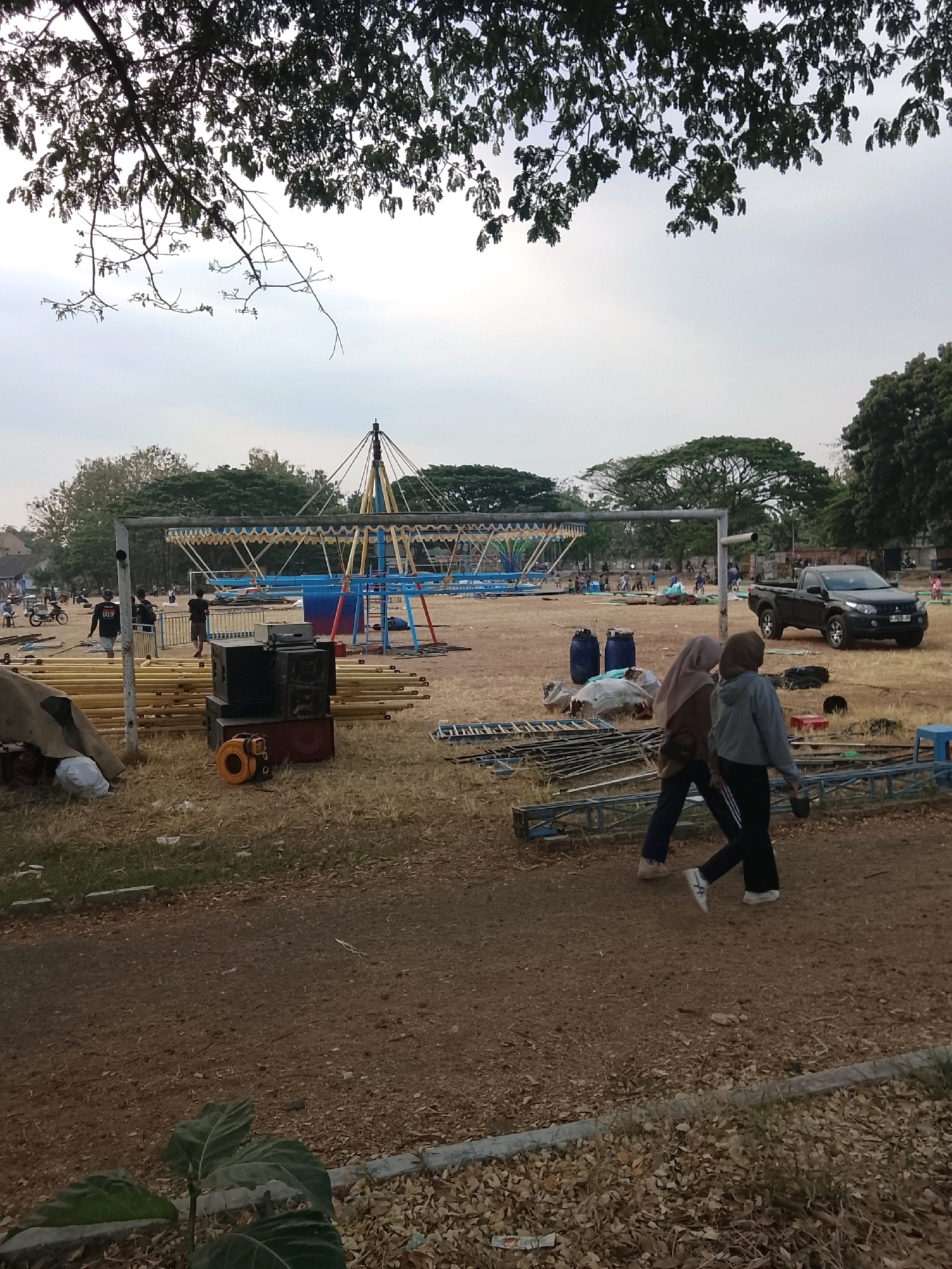 The image depicts an outdoor scene in Ujungrusi, Indonesia, likely a park or open field. In the foreground, two women are walking from right to left, both wearing hijabs and casual attire. One woman is wearing a gray hoodie and black pants, while the other is in a brown hijab and dark pants. Several stacks of yellow poles and metal scaffolding are visible, suggesting ongoing construction or setup for an event. A black pickup truck is parked in the middle ground to the right. Further back, a large, skeletal structure that appears to be a carousel or similar amusement ride is partially assembled. Various other individuals, some on motorcycles, are scattered throughout the area, and trees form the backdrop under a hazy sky.