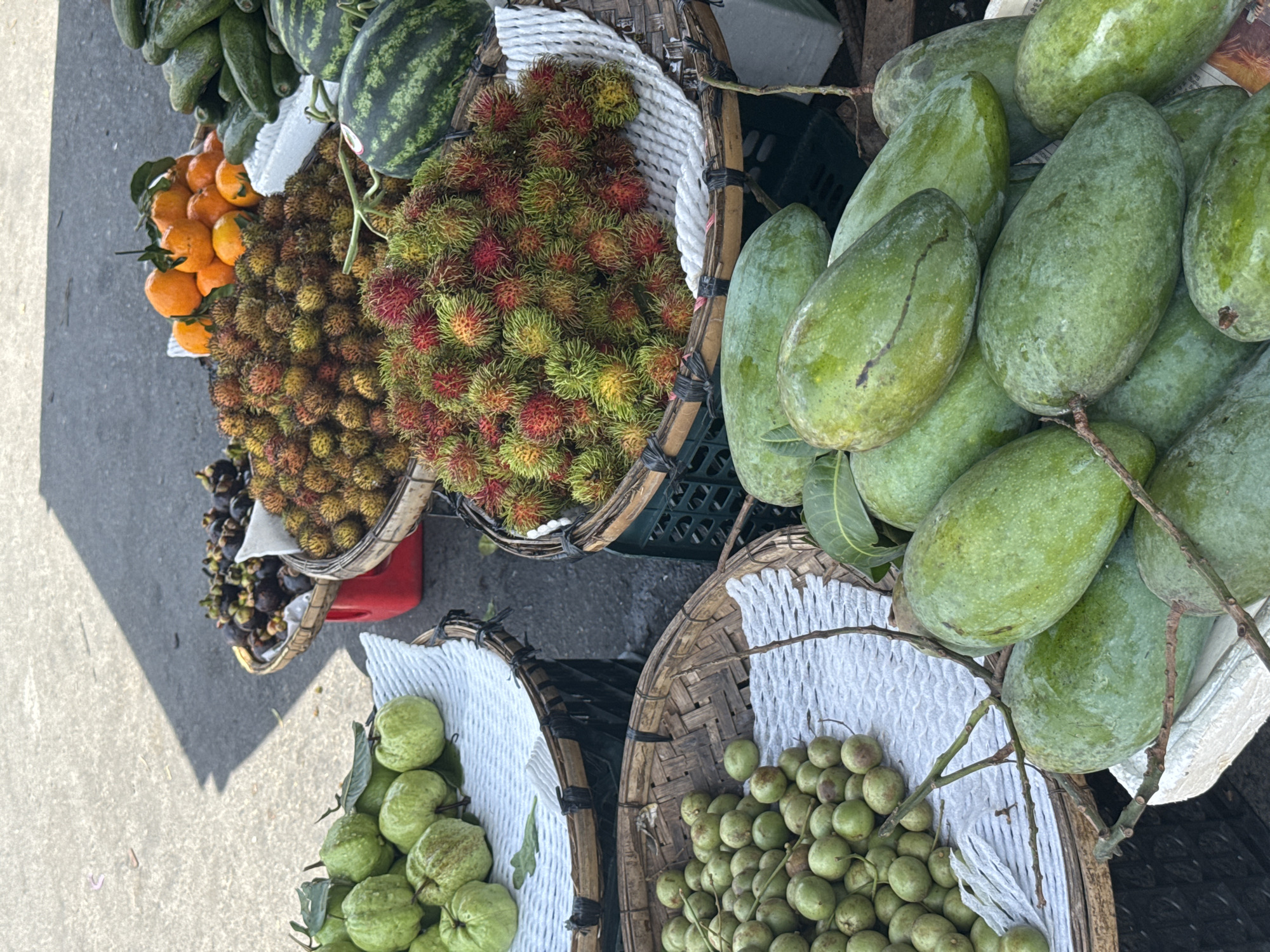 A fruit stand in Nha Trang Ward, Vietnam, displays a variety of fresh fruits. In the foreground, a pile of green mangoes rests on a white surface. To the left, a basket overflows with small, round green fruits, possibly grapes or a similar berry. Behind this, another basket holds larger, oval-shaped green fruits, likely guavas. Further back, a basket filled with rambutan, a tropical fruit with spiky red and green skin, is visible. Next to the rambutan, a pile of mangosteens, dark purple fruits with a crown-like top, sits in a woven basket. To the right of the mangosteens, a cluster of bright orange fruits, likely tangerines, are neatly arranged. In the background, parts of watermelons and possibly avocados are visible, suggesting a diverse selection of produce. The fruits are laid out on a paved surface, possibly a street or market walkway, with a shadow cast from an unseen structure. The scene captures a typical open-air market in Vietnam.