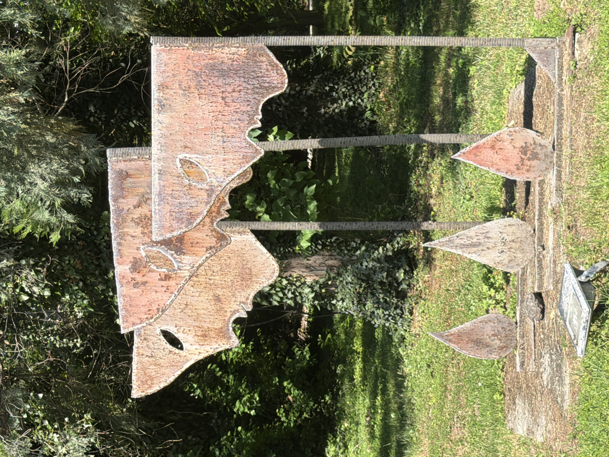 The image shows an outdoor sculpture located in a park or garden setting in Pazardzhik, Bulgaria. The sculpture is made of weathered metal and consists of two abstract, flag-like shapes at the top, supported by vertical metal poles. The shapes have irregular edges and appear to be oxidized, with rust coloring their surfaces. Cut-out leaf or eye shapes are visible on the upper sections. In front of the main sculpture, three tear-drop shaped metal pieces are embedded in a concrete base. The ground around the sculpture is covered in grass and some foliage. In the foreground, a small, flat, rectangular object, possibly a light fixture, is situated on the grass. The background is filled with lush green trees and vegetation, suggesting a natural and somewhat overgrown environment. The lighting indicates it is daytime, likely sunny, casting some shadows.