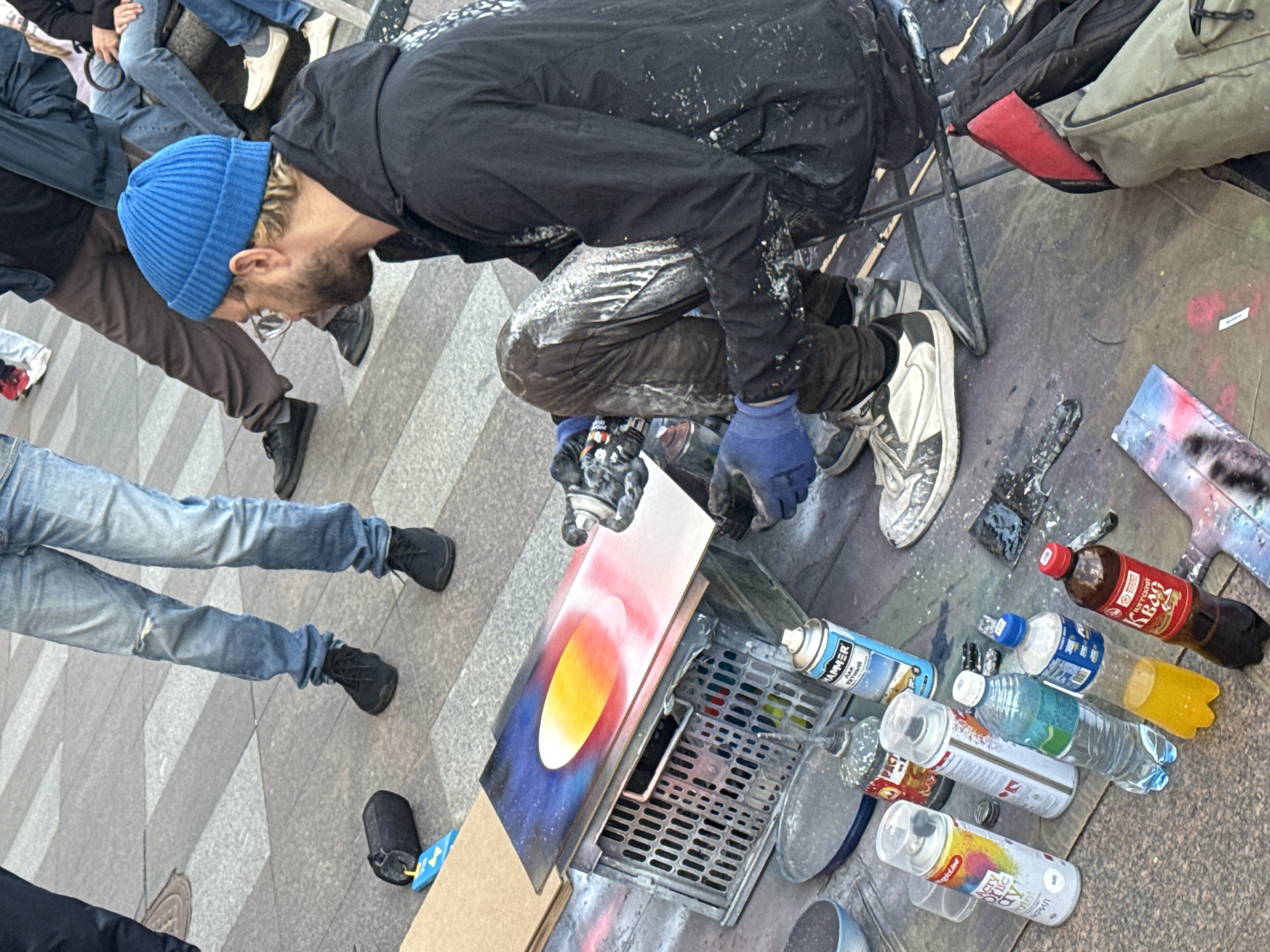 An artist is spray-painting an abstract cosmic scene of a planet with rings onto a board. He is wearing a blue beanie, clear glasses, a black jacket and pants splattered with paint, and blue gloves. The board is propped up on a grey plastic crate on the ground. Various spray cans and bottles are scattered around the artist. In the background, other people are sitting and standing, some also in casual clothing. The scene appears to be set in a public urban area, possibly a plaza or street, with patterned paving stones visible. The overall lighting suggests an overcast day.