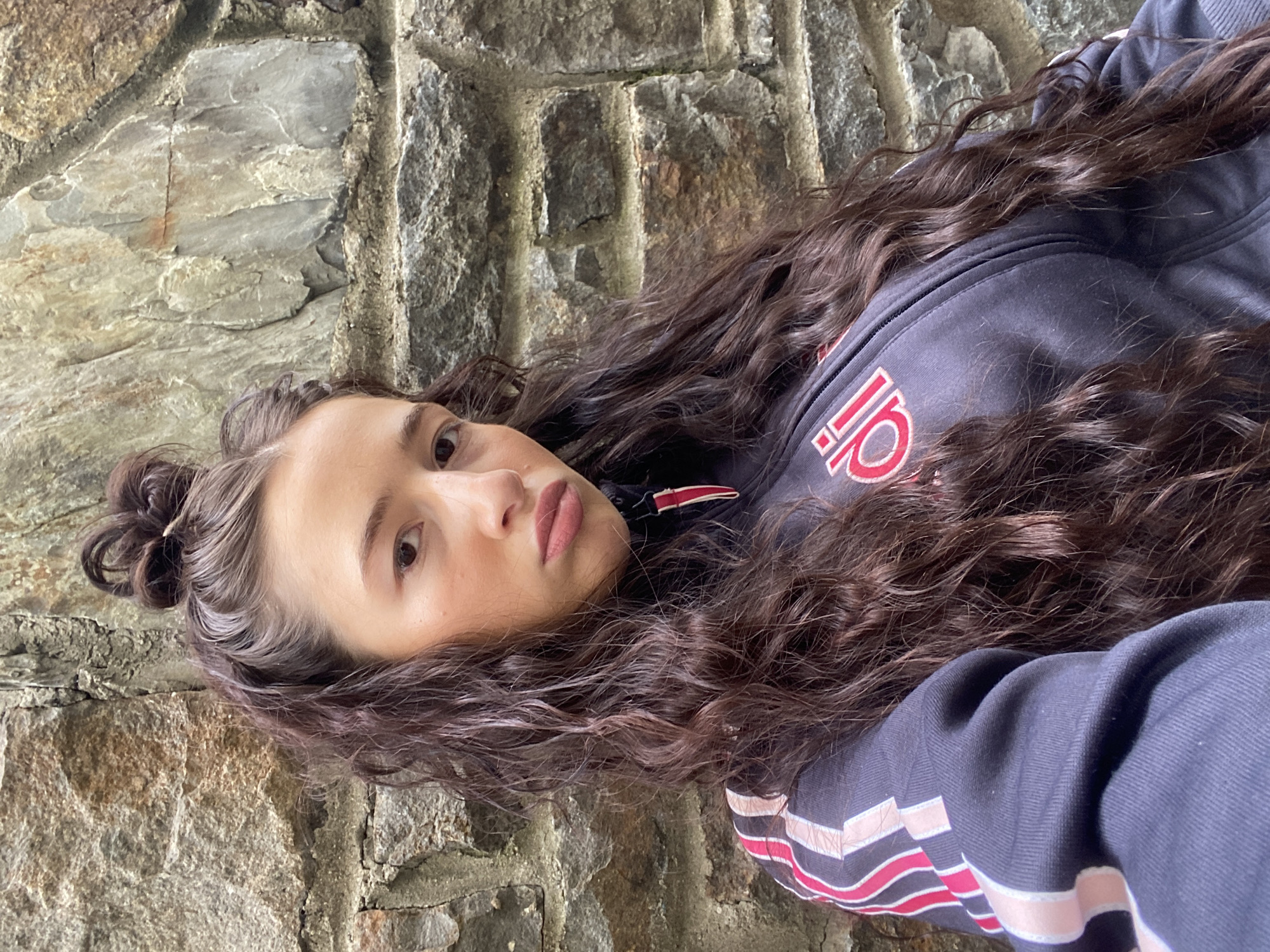 A young woman with long, dark curly hair lies against a stone wall. Her hair is styled with a small bun on the top of her head. She is wearing a dark blue hooded sweatshirt with red and white stripes and a red and white logo. The stone wall behind her is composed of various-sized grey stones. The location appears to be outdoors, but there are no other obvious environmental clues to specifically place the scene in Llan Ffestiniog, United Kingdom.