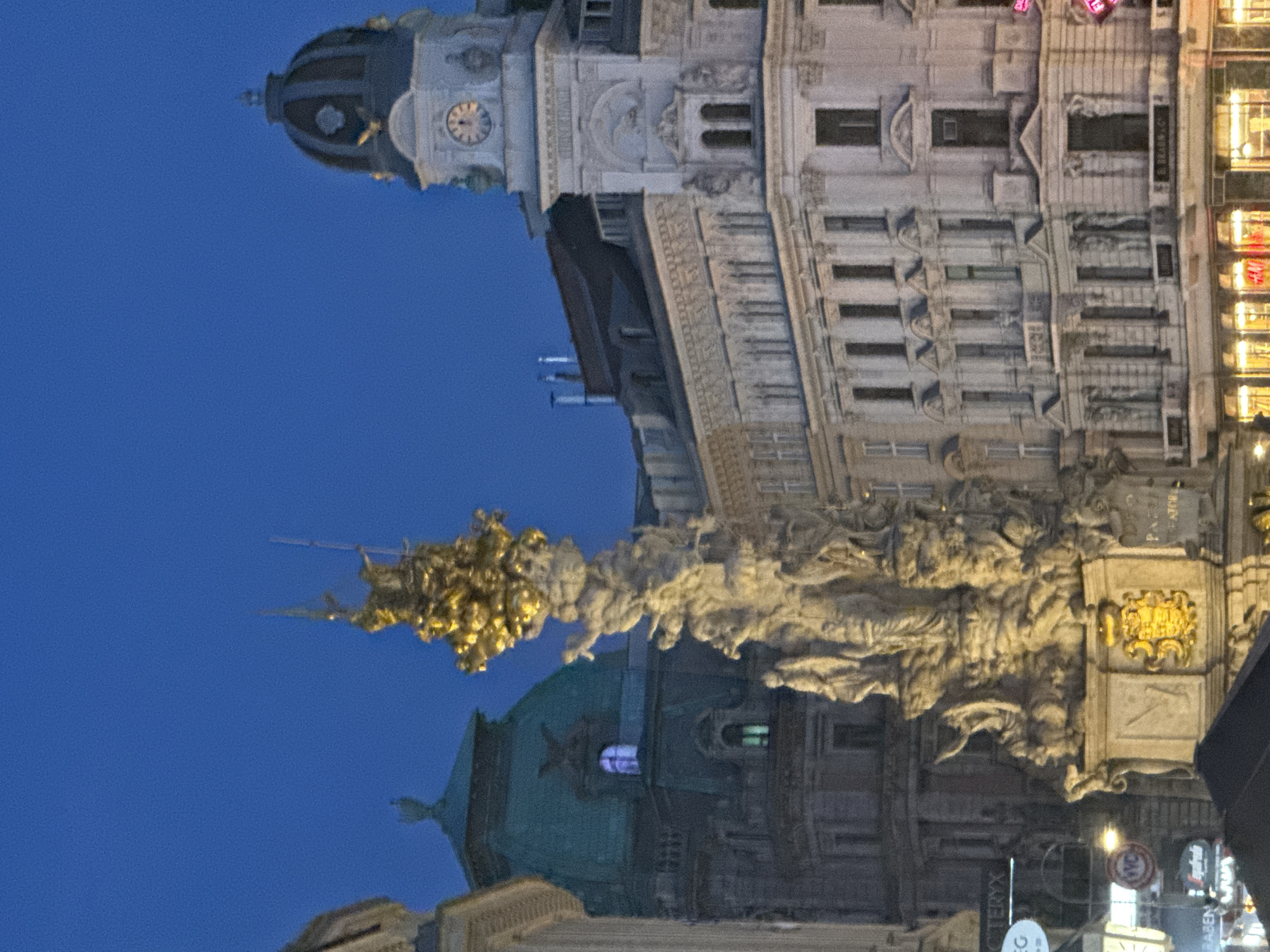 The image shows a nighttime view of the Plague Column (Pestsäule) in Vienna, Austria. The monument, made of stone and topped with a golden sculpture, is illuminated from below, casting a glow against the dark blue twilight sky. To the right of the Plague Column, a portion of a grand white building with an ornate facade and a domed clock tower is visible. The building features multiple windows, arched doorways, and decorative sculptures. To the left of the Plague Column, another building with a green, domed roof is partially visible. In the foreground, lower-lying structures with lit windows and signage are present, suggesting a commercial area. The overall scene depicts an urban setting at dusk, highlighting architectural landmarks and the ambient lighting of the city.