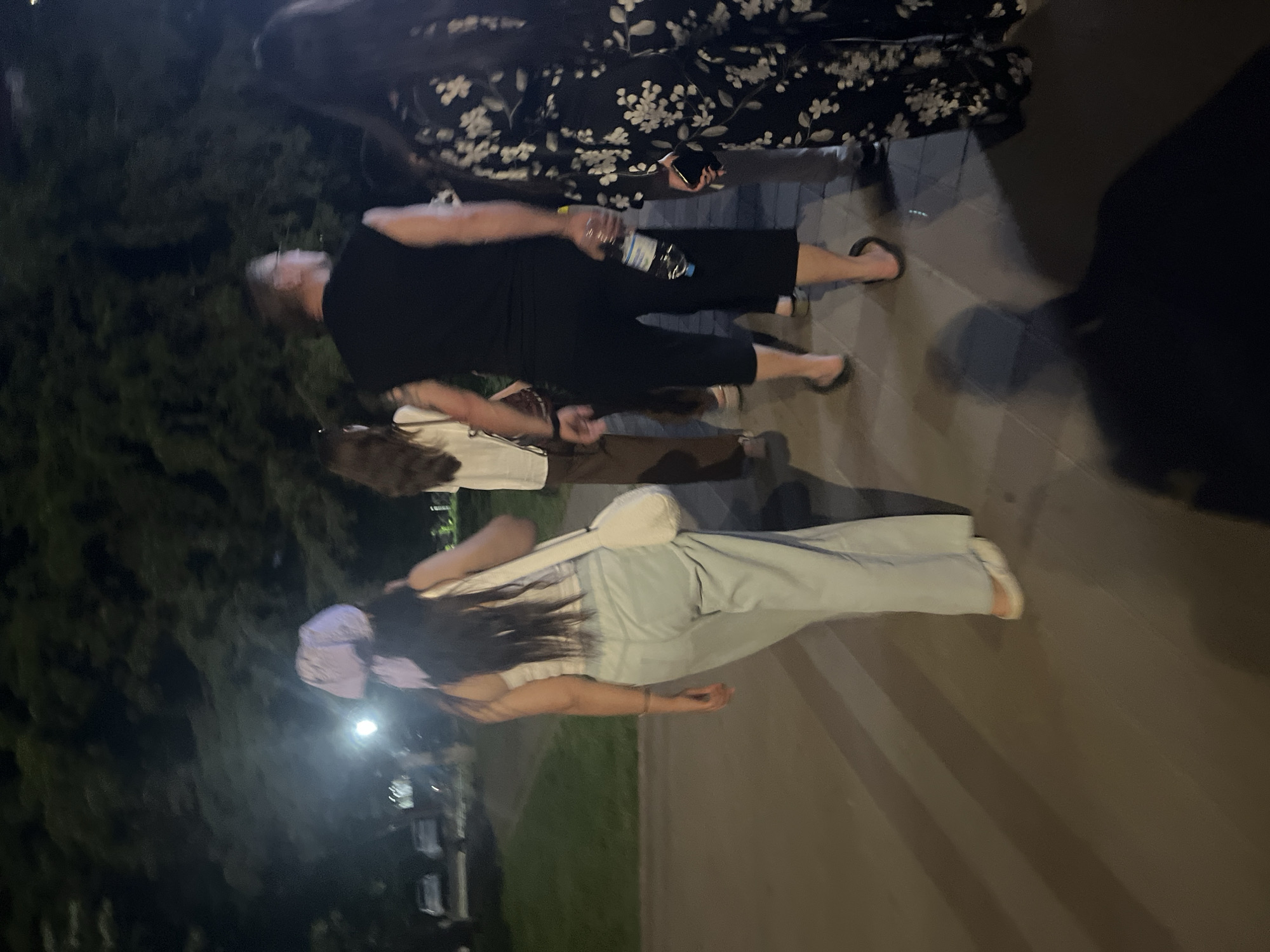 The image depicts a group of people walking on a paved walkway at night. The scene appears to be outdoors, with trees in the background and artificial lighting casting shadows. A woman in the foreground is walking away from the camera, wearing a light-colored tank top, pale blue wide-leg pants, and a light purple headscarf. She is carrying a white purse. To her right, a man in a black sleeveless shirt and black capri pants is walking, holding a bottle of water. Behind him, a woman in a white top and brown pants is visible. Further to the right, another woman is walking in a black floral patterned jumpsuit. Cars are parked in the background on the left side of the image. The overall impression is of a casual evening stroll in an urban or park-like setting.