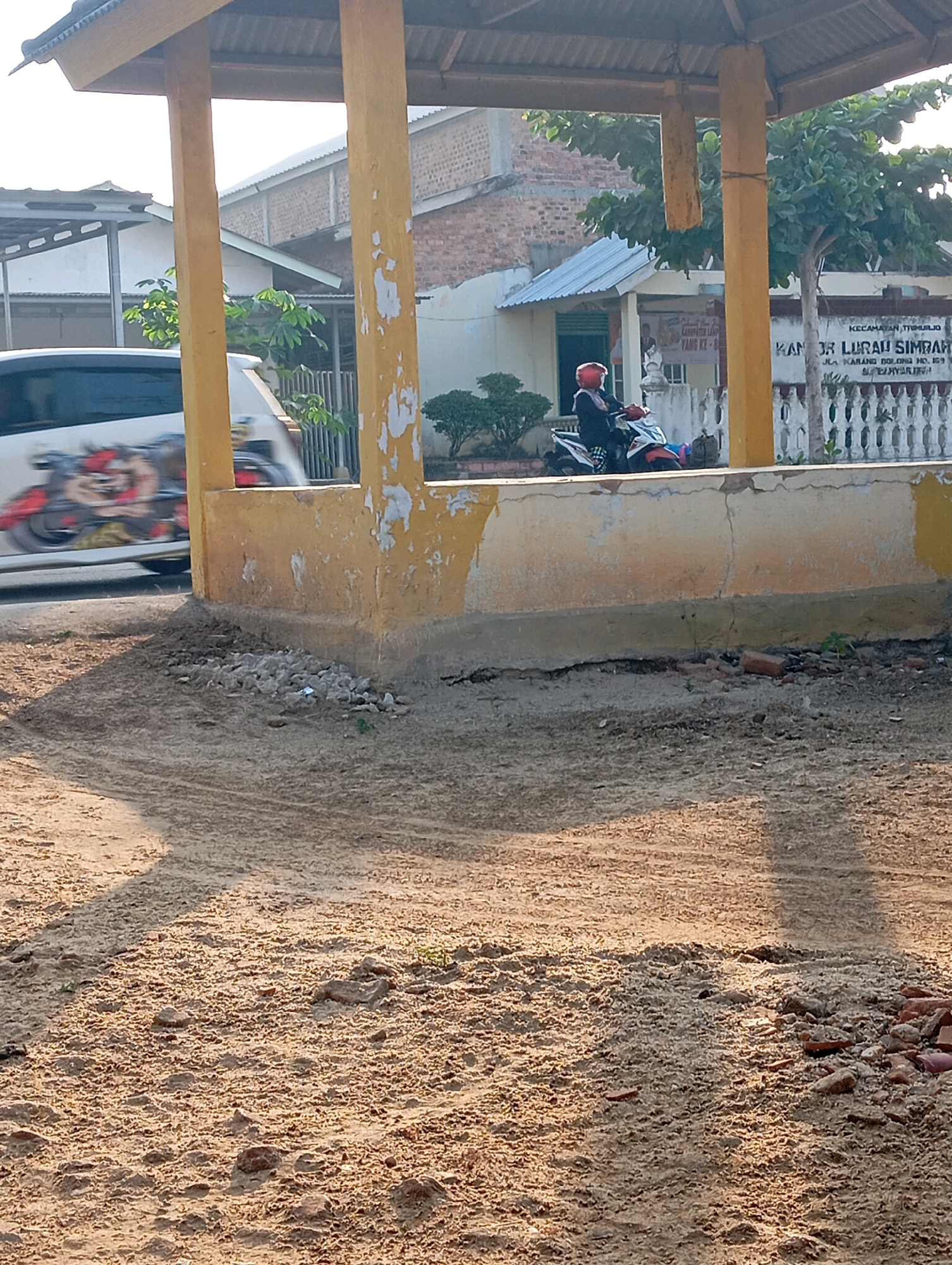 The image depicts an outdoor scene in Adi Puro, Indonesia, characterized by a sandy, uneven ground in the foreground and a partially covered structure with yellow pillars and a corrugated metal roof. Behind the structure, a building with a light-colored facade and a tiled roof is visible, along with a white fence and vegetation. A white van with graphic art is partially seen on the left side of the frame, and a person wearing a red helmet is seated on a motorcycle to the right of the van. A sign with Indonesian text is mounted on the fence. The lighting suggests it is daytime, with shadows cast on the ground.