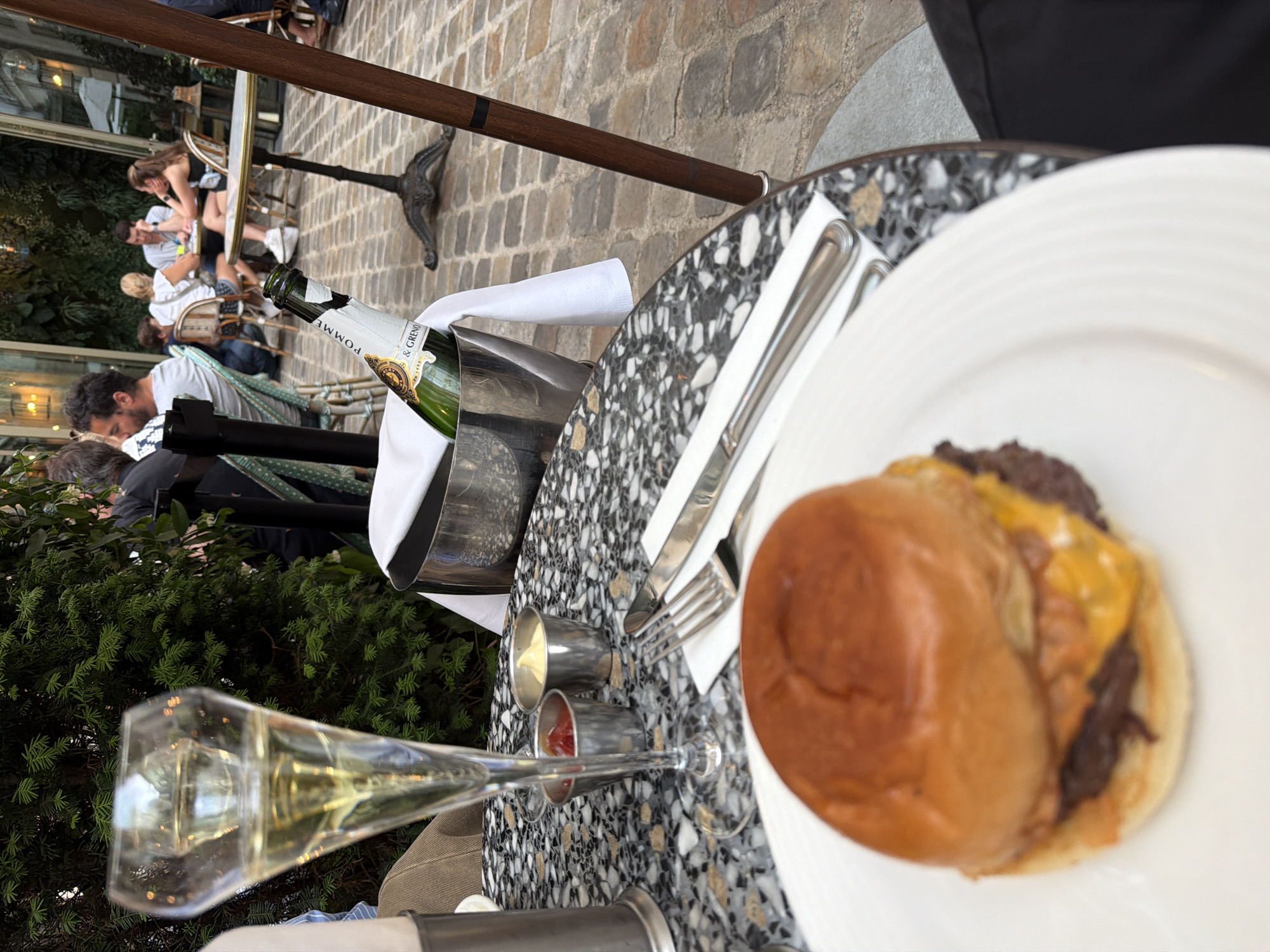 The scene depicts a table at a restaurant in Paris, France. A burger sits on a white plate, next to silverware and small containers, possibly holding condiments. A champagne glass filled with bubbly rests near an ice bucket containing a bottle of Pommery champagne. People are seated at nearby tables. The table has a speckled black and white surface, and the ground is paved with cobblestones. A green hedge is visible in the background.