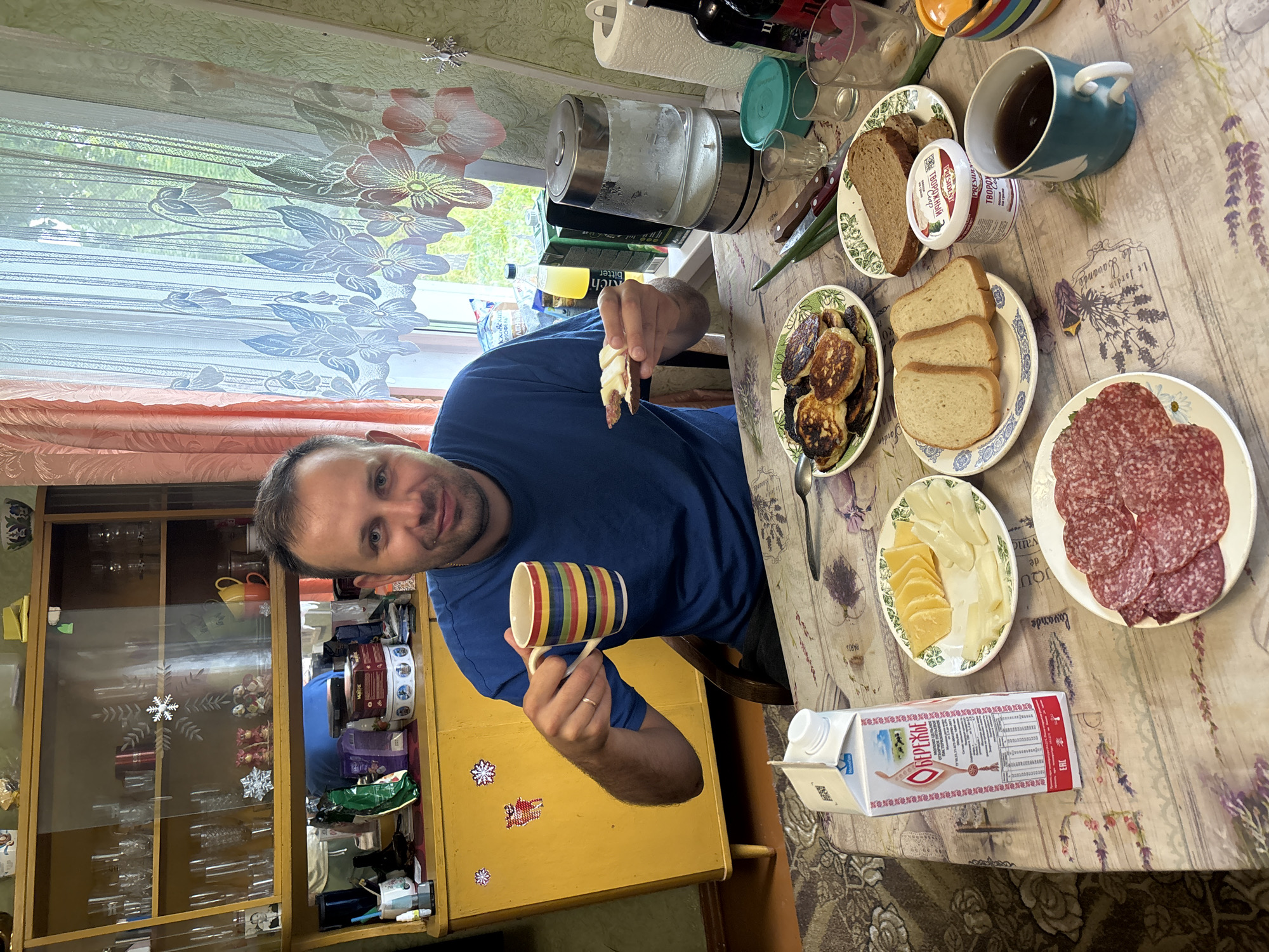 A man is sitting at a table in a dining room in Cherkasovo, Russia, holding a mug and preparing to eat. The man is wearing a blue t-shirt and has short brown hair. He is holding a colorful mug in his right hand and a piece of bread with butter in his left hand. On the table in front of him are plates of food, including pancakes, cheese, sliced salami, and bread. There is also a carton of milk, a jar of yogurt, glasses, and a tea cup. In the background, a cabinet filled with glassware and a window with curtains can be seen. The room appears to be well-lit and domestic.