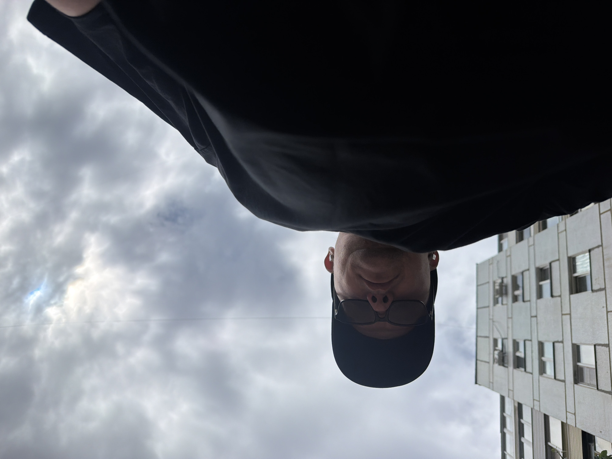 This is a low-angle, upside-down selfie taken outdoors in Chaikovsky, Russia. The subject is a person wearing a black baseball cap, sunglasses, and a black t-shirt. They are looking directly at the camera. Behind them, a modern, multi-story building with many windows is visible, constructed of light-colored concrete panels. The sky is overcast with heavy, grey clouds, suggesting a dull or possibly rainy day. The lighting is diffused due to the cloud cover, indicating it is likely daytime. There is no visible text in the image.
