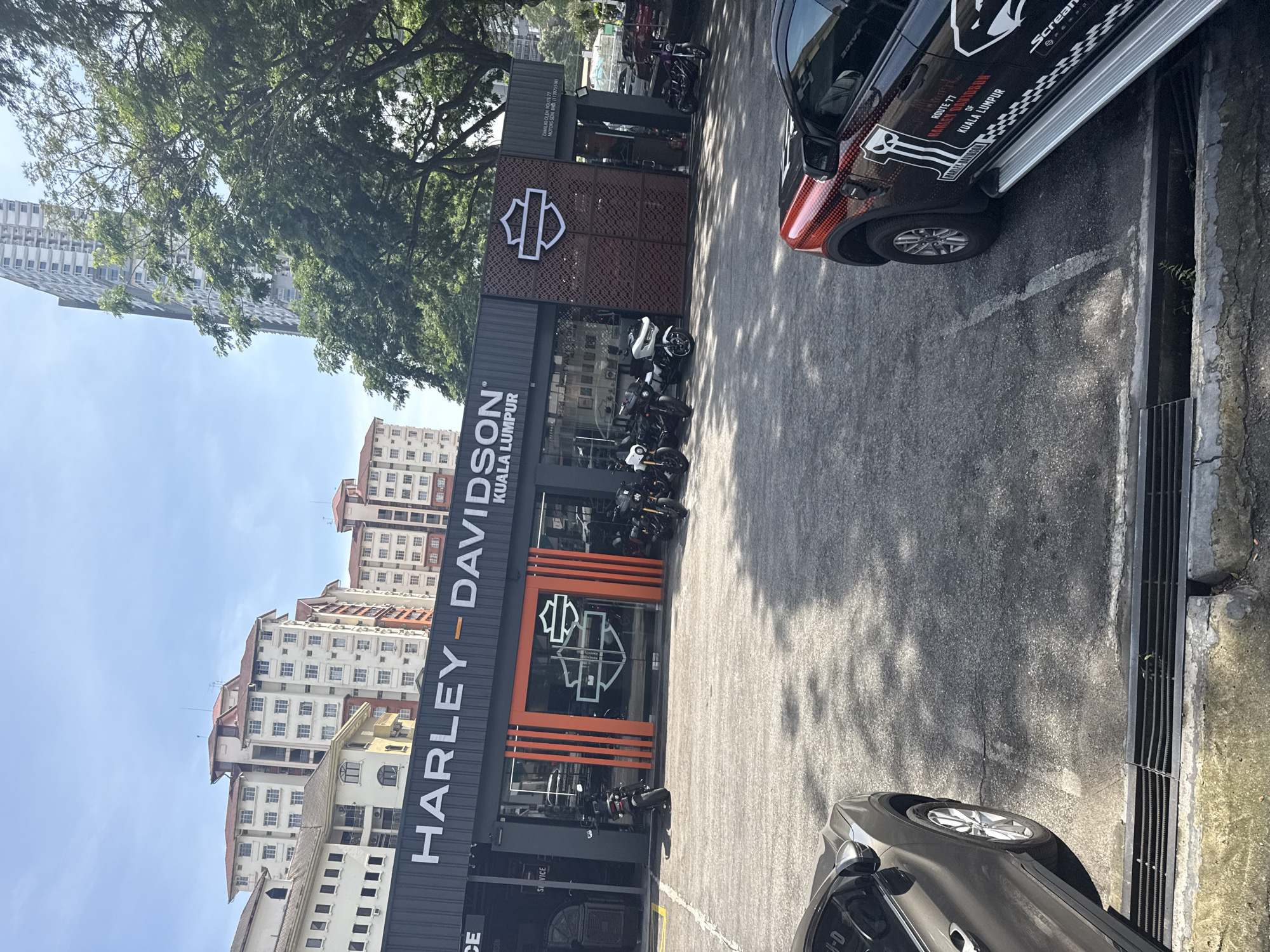 The image shows the Harley-Davidson dealership in Kuala Lumpur, Malaysia. The dealership's sign, "HARLEY-DAVIDSON KUALA LUMPUR", is prominently displayed on the building's facade. Several motorcycles are parked in front of the dealership. A black car with Harley-Davidson branding is parked to the right. A grey sedan is partially visible at the bottom of the image. Apartment buildings are visible in the background. The sky is partially visible with tree foliage obscuring some of it. The scene is taken in daylight.