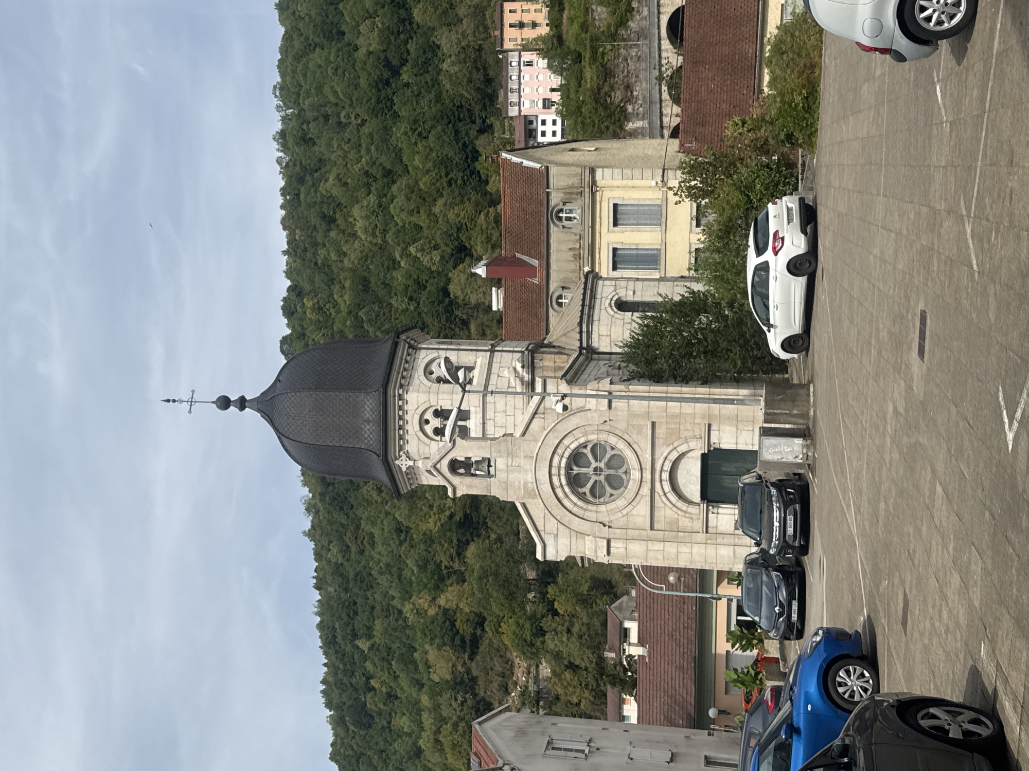 A wide shot captures a church with a dark dome and a pale stone facade, featuring a rose window and a bell tower, situated in the town of Saint-Claude, France. The church is positioned in the foreground of a verdant, tree-covered hill. In front of the church, several cars are parked in a paved lot. These include a blue hatchback, a black sedan, a black SUV, and a white hatchback. Behind the church, a row of buildings with red tile roofs and colorful facades is visible, climbing the hillside. The sky above is overcast with a few wisps of clouds.