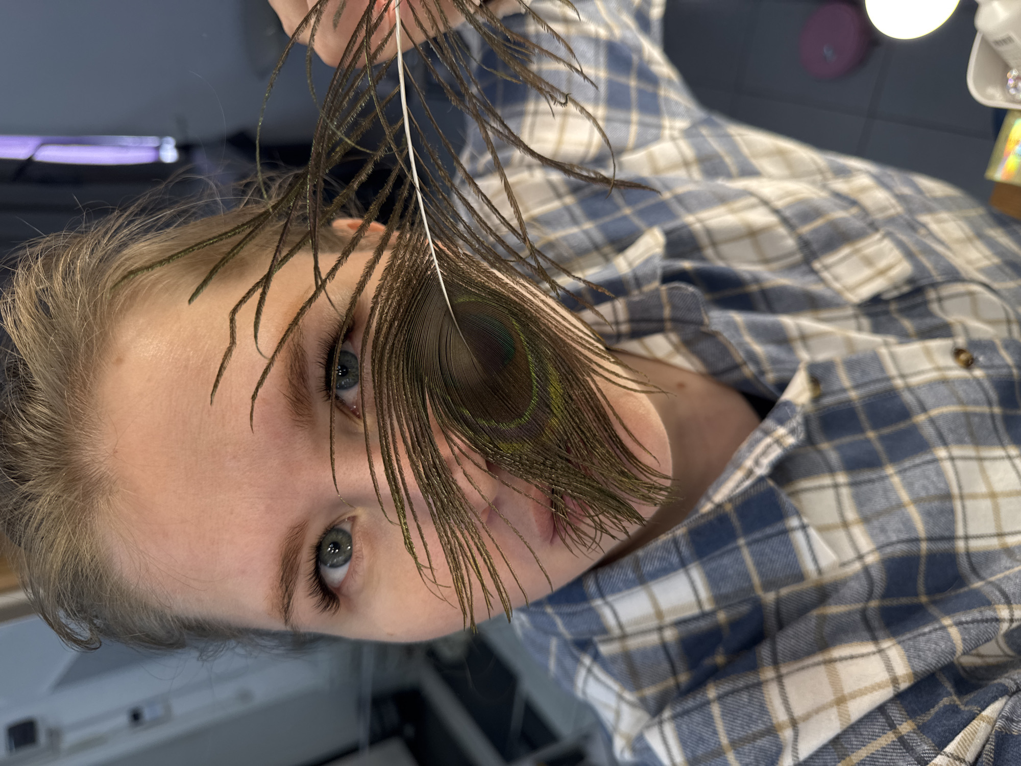 A fair-skinned person is holding a peacock feather in front of their face. The person is wearing a blue and white checkered shirt. The person's hair is blonde and pulled back. The eyes are light blue. The location is not discernible as Perm, Russia based on the image.