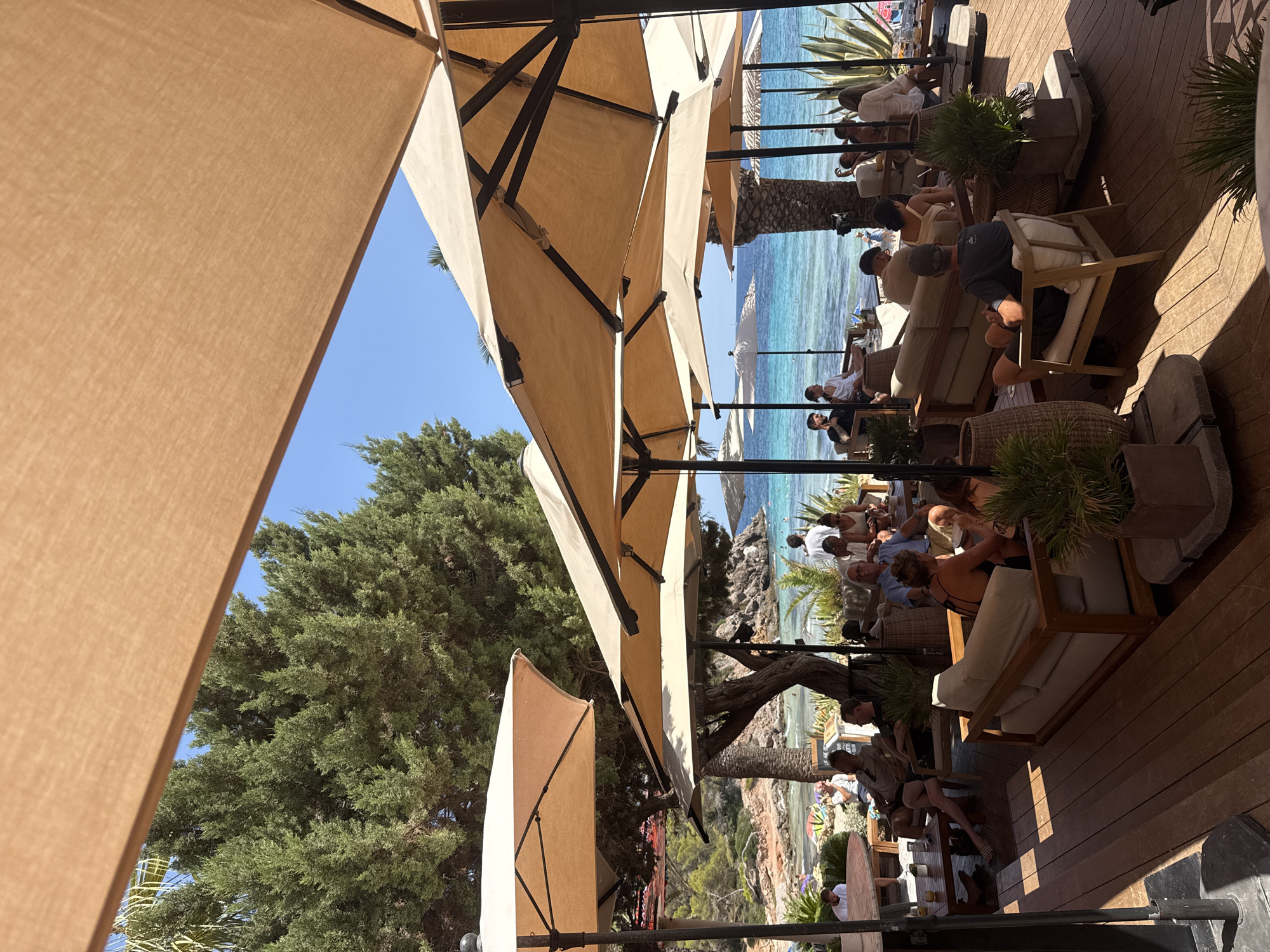 A group of people are seated at an outdoor dining area with a view of the ocean. The scene is located in Santa Eulària des Riu, Spain. Several large, tan umbrellas provide shade. In the foreground, a man with a gray t-shirt and black cap is sitting in a chair. To his left, a couch with white cushions is occupied by a man and woman. To the left of the couch, a round table has two men seated and drinks on it. In the middle ground, more people are seated at tables and couches, some facing the ocean. Beyond the seating area, a beach with people can be seen, and the turquoise water stretches to the horizon. Lush green trees and plants are visible throughout the scene. The ground is made of wooden planks.