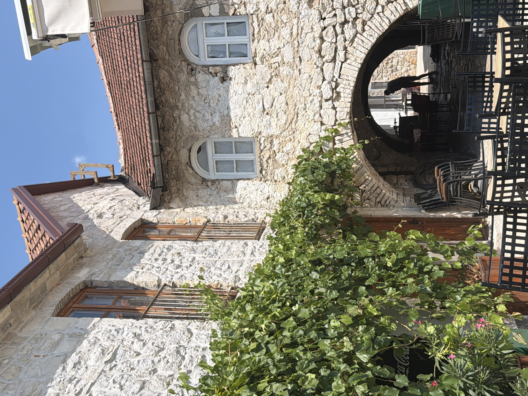 In Trebinje, Bosnia and Herzegovina, an outdoor seating area of a cafe is depicted. The cafe is situated within a stone building with a red-tiled roof. Lush green ivy cascades down the stone facade, partially obscuring a wooden door and a Coca-Cola advertisement. Arched stone passageways lead into the interior of the building, where tables and chairs are visible. A person, silhouetted against the light, is sweeping the floor within the arched entryway. Stacked chairs and tables are arranged in the foreground and around the seating area. The scene suggests a warm, sunny day, with a clear blue sky overhead.