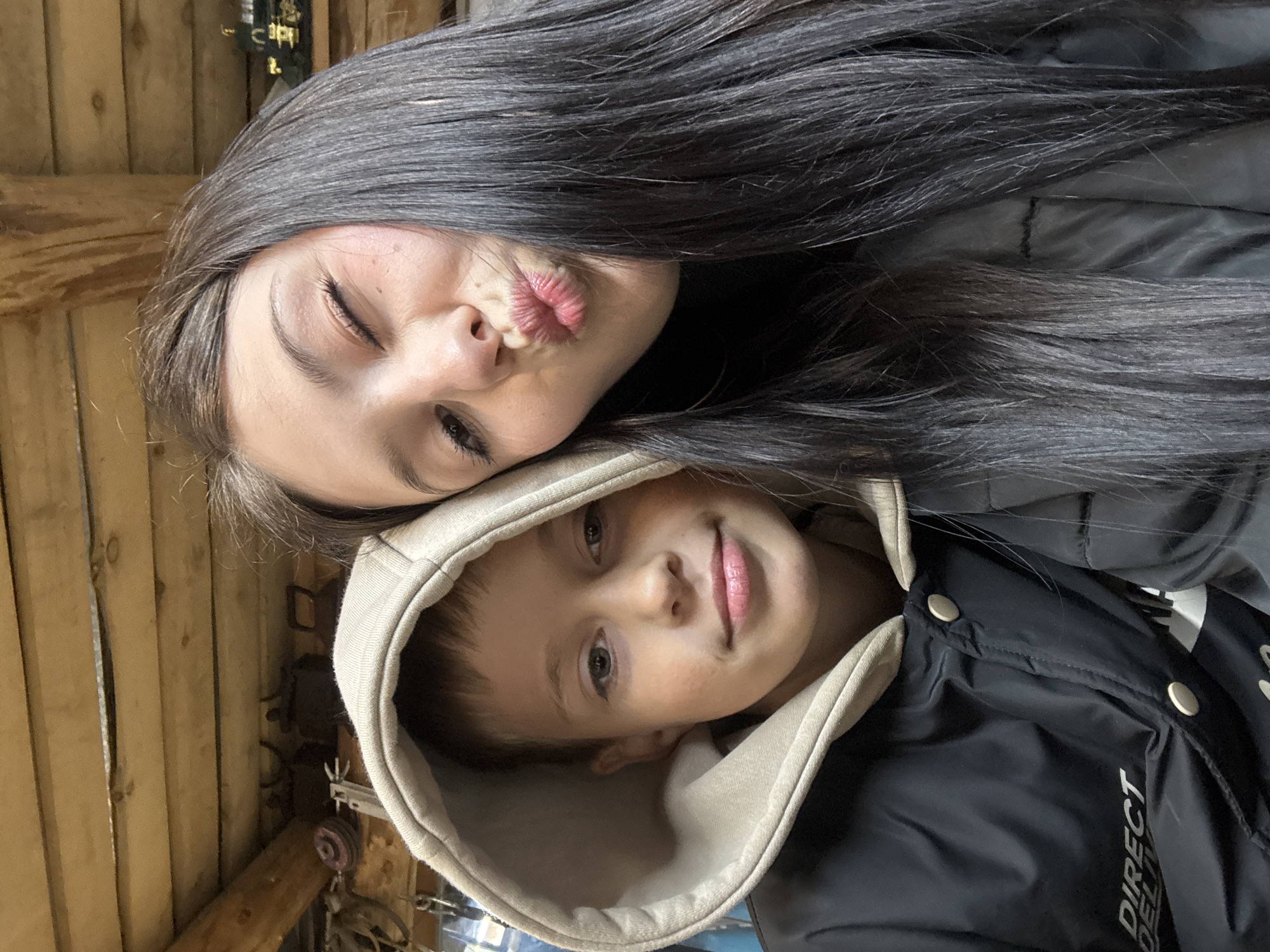 A young woman with dark hair is making a kissing face with her left eye closed, while a young boy wearing a hooded jacket looks at the camera. They appear to be in a wood shed or barn. The location is Фоки, Russia.