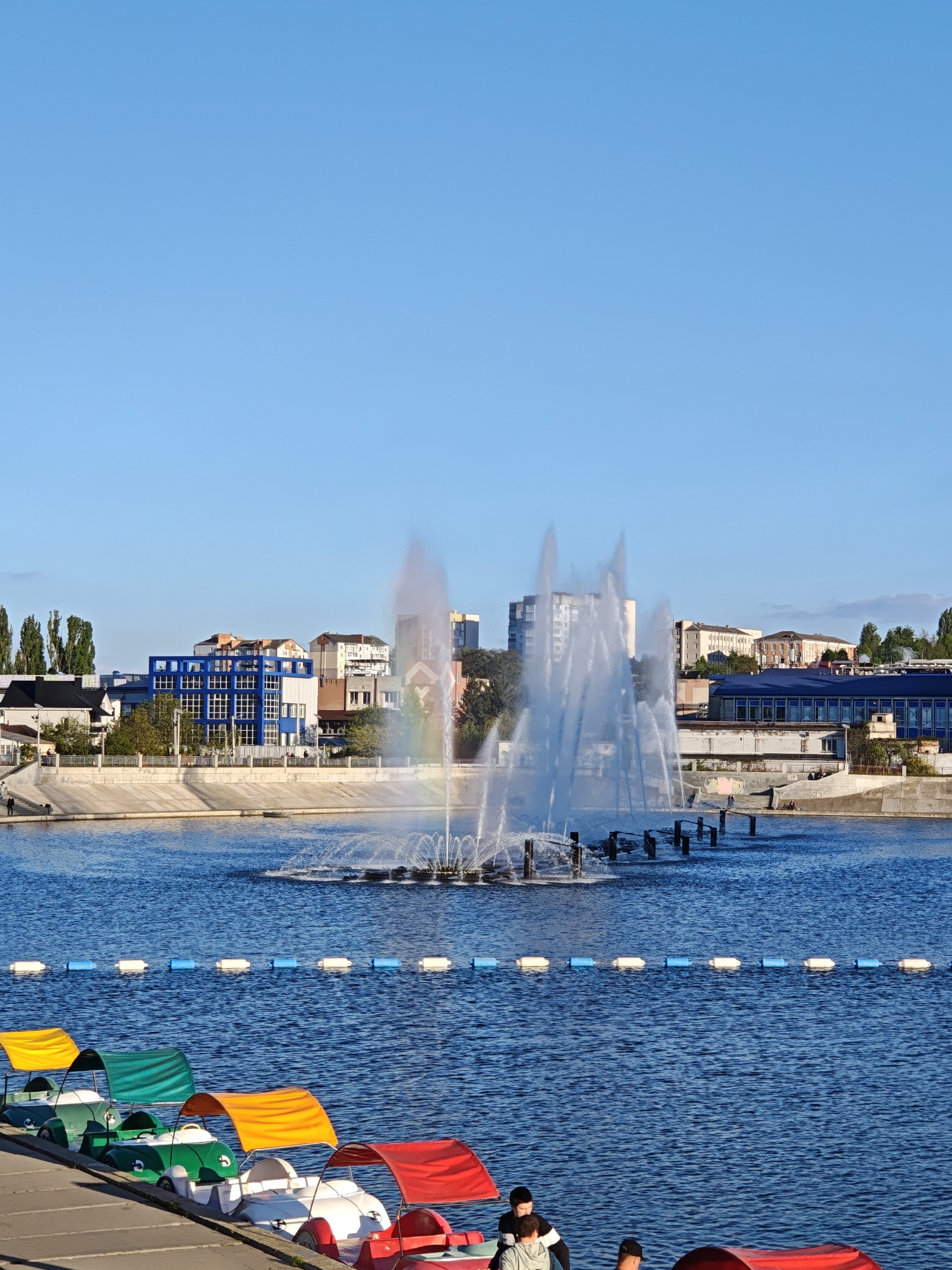 The image shows a waterscape in Vinnytsia, Ukraine. In the background is a fountain with a partial rainbow, and behind the fountain are buildings and trees. In the foreground, several colorful swan-shaped paddle boats are docked along the shore of a body of water. The paddle boats are covered in red, orange, green, and yellow awnings. Buoys are visible in the water separating the foreground boats from the background fountain. In the lower right, people appear to be near the boats. The sky is clear and blue.