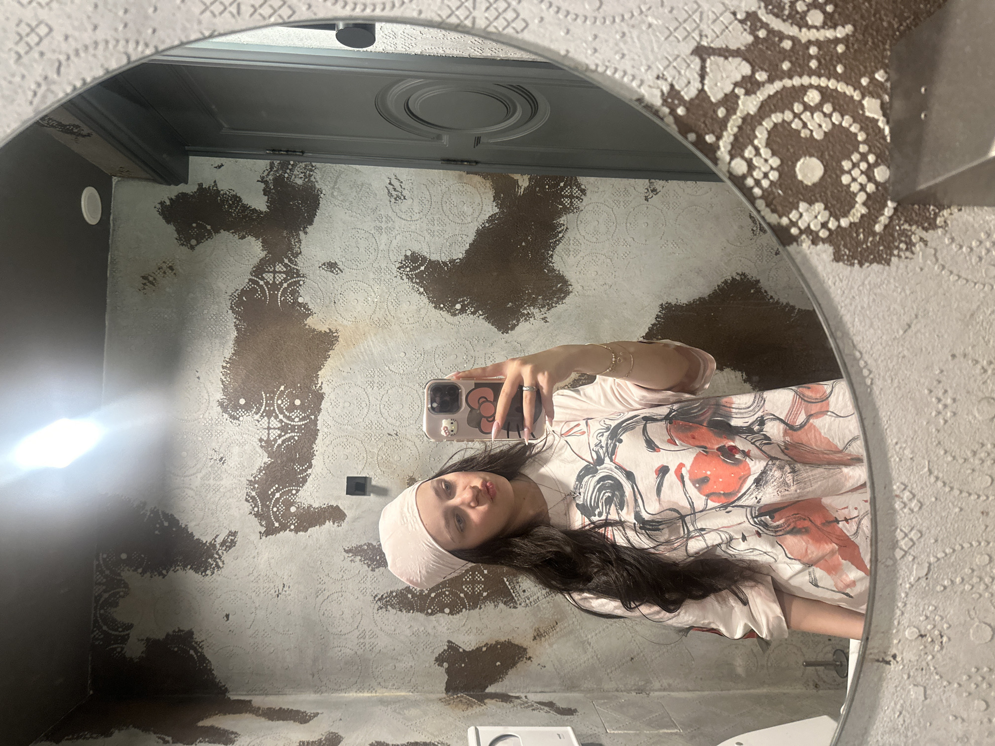 A young woman with dark hair and wearing a headscarf and a patterned top takes a selfie in a mirror. The mirror is round and has a textured border. The background shows a wall with peeling wallpaper in brown and white patterns, revealing what looks like a layer of patterned wallpaper underneath. A light fixture is visible on the ceiling. No information allows inference on the city of Yerevan, Armenia.