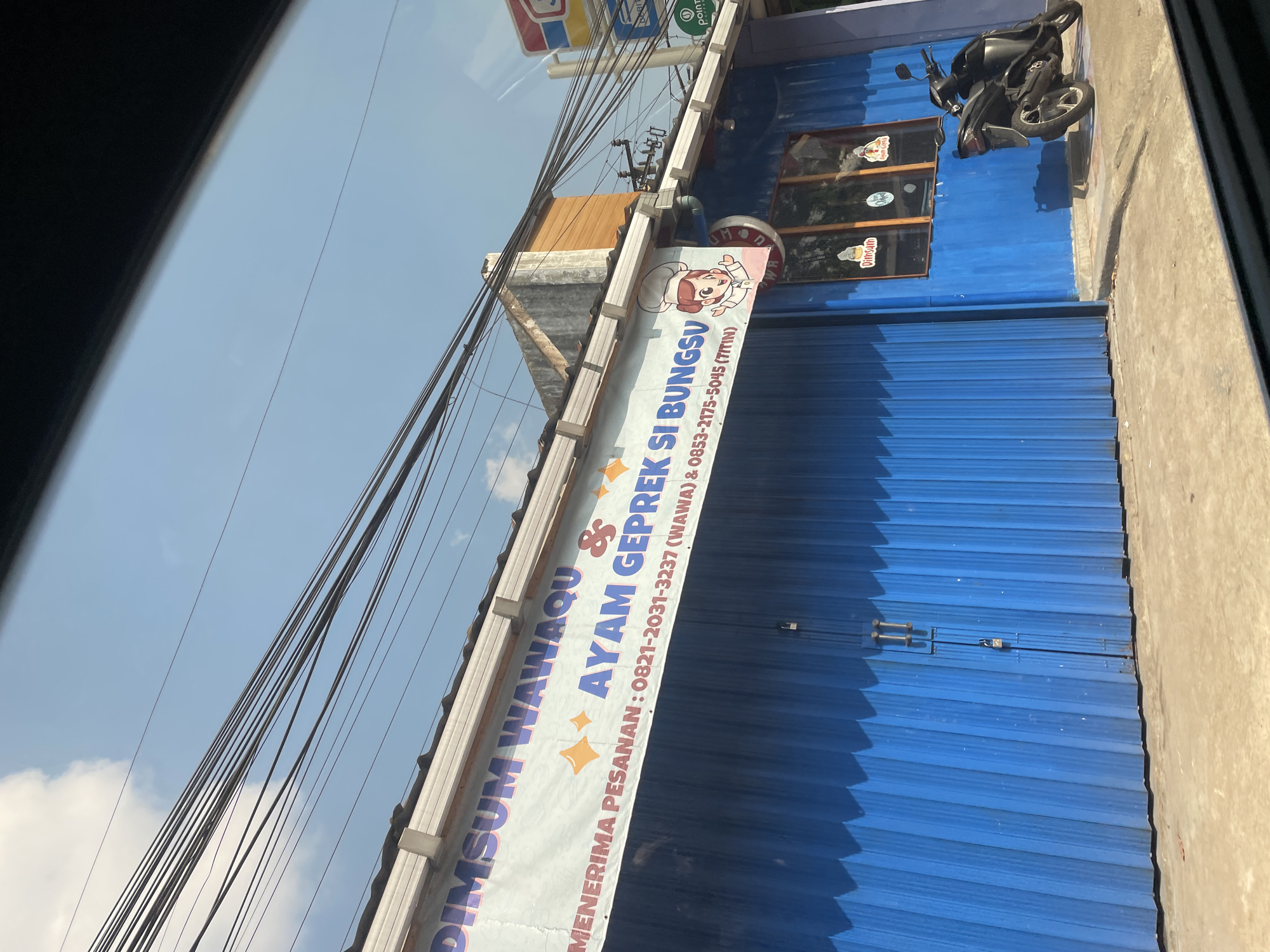 The image displays the exterior of a bright blue commercial building, identified by a white banner as "DIMSUM WAWAQU & AYAM GEPREK SI BUNGSUN" (Dimsum Wawaqu & Fried Chicken Si Bungsu). The banner also states "MENERIMA PESANAN :" (Receiving orders :) followed by two phone numbers: "0821-2031-3237 (WAWA)" and "0853-2175-5045 (TITIN)". A cartoon chef character is depicted on the right side of the banner. The building's front features a prominent blue corrugated metal section on the left, which is closed and secured by a padlock and two vertical metal bars. To the right, there is a smooth blue wall with three small, rectangular windows featuring wooden frames. Stickers, one appearing to say "Dimsum," are visible on these windows. A dark-colored scooter is parked on the light-colored concrete surface in front of the right side of the building. Above the building, numerous black electrical wires crisscross against a clear blue sky with sparse white clouds. Shadows indicate direct sunlight. On the far right, partial views of other commercial signage, including a red and white sign (possibly Indomaret) and a green "Point" logo, are visible, along with parts of a utility pole structure and additional wires. The scene is located in Ciater, Indonesia. No people are visible.
