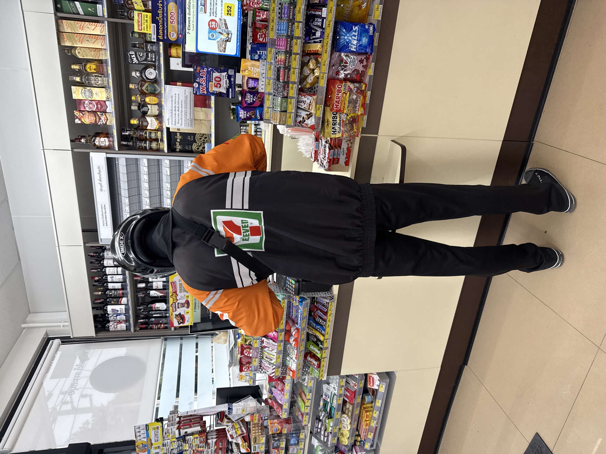 A person wearing a black and orange 7-Eleven uniform and a black helmet stands at a checkout counter in a 7-Eleven convenience store, facing away from the camera. The person is holding a payment device. The counter is lined with various snacks and beverages. Behind the counter, shelves are stocked with more merchandise, including bottles of alcohol and packaged goods. The floor is tiled in a neutral tone. The store appears to be in Ko Samui District, Thailand.