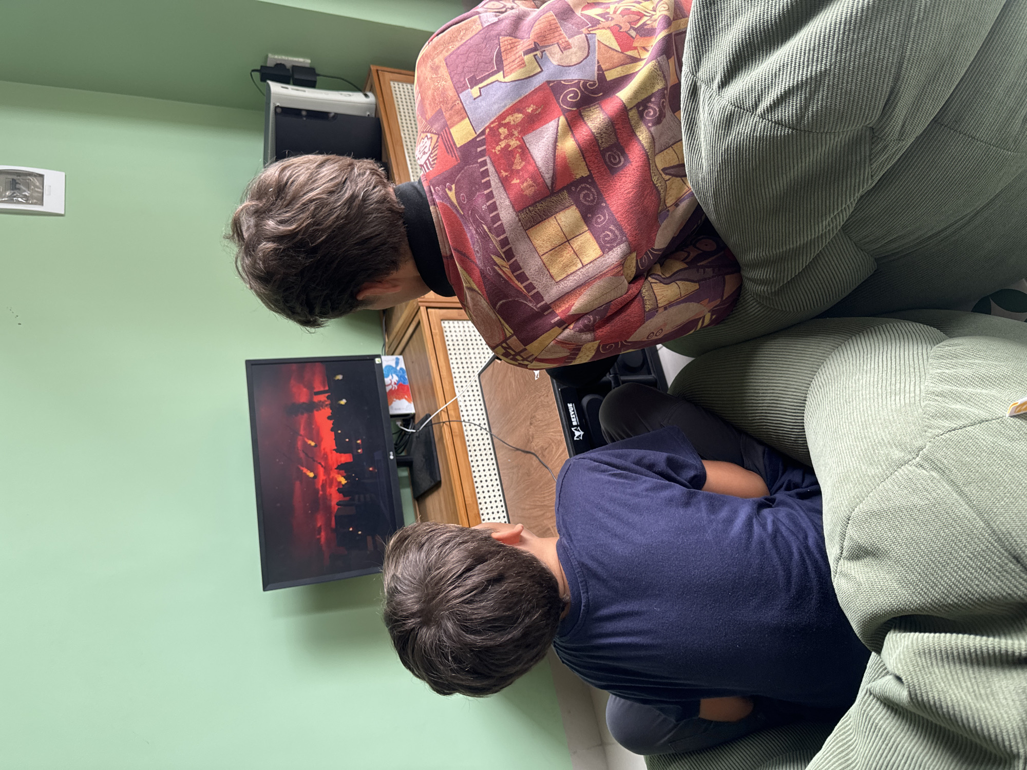 Two people are looking at a computer screen displaying an image of a city burning. One person is wearing a red and tan patterned shirt and green corduroy pants. The other person is wearing a dark blue shirt and grey pants. The location may be in Phu Thuy Ward, Vietnam. The room has a light green wall. There is a cabinet next to the computer, and a light switch on the wall.