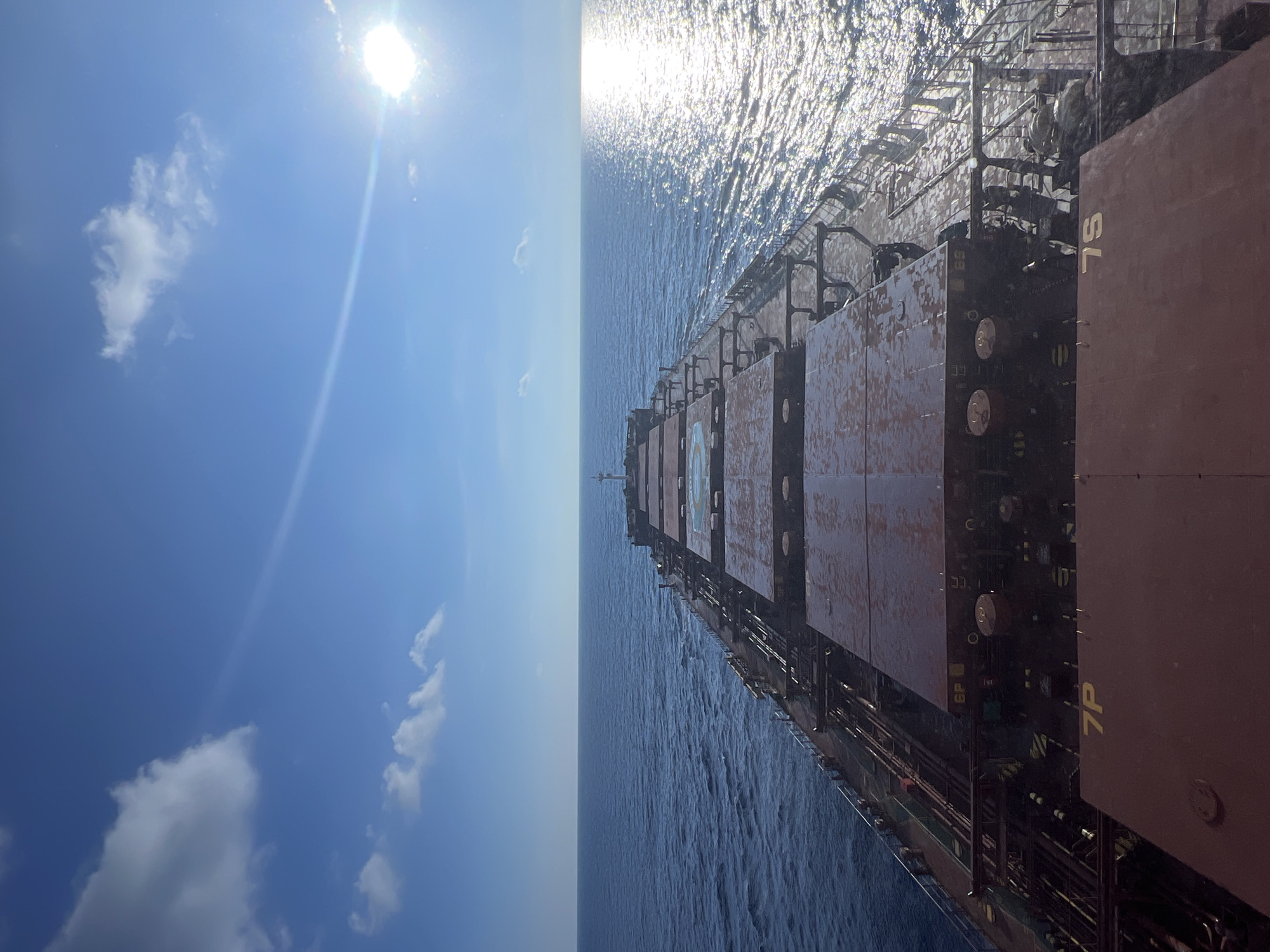 An aerial shot looks down a long, flat-topped cargo ship sailing on a calm, blue ocean under a bright, sunny sky. The sun glints off the water to the right, creating a shimmering effect. A few white clouds are scattered across the sky. The ship's deck is covered in large, reddish-brown hatches, some marked with yellow lettering like "7P" and "7S". Pipes and equipment are visible between the hatches. The ship appears to be moving forward, with the bow extending into the distance. The horizon is sharp, separating the sea from the sky.